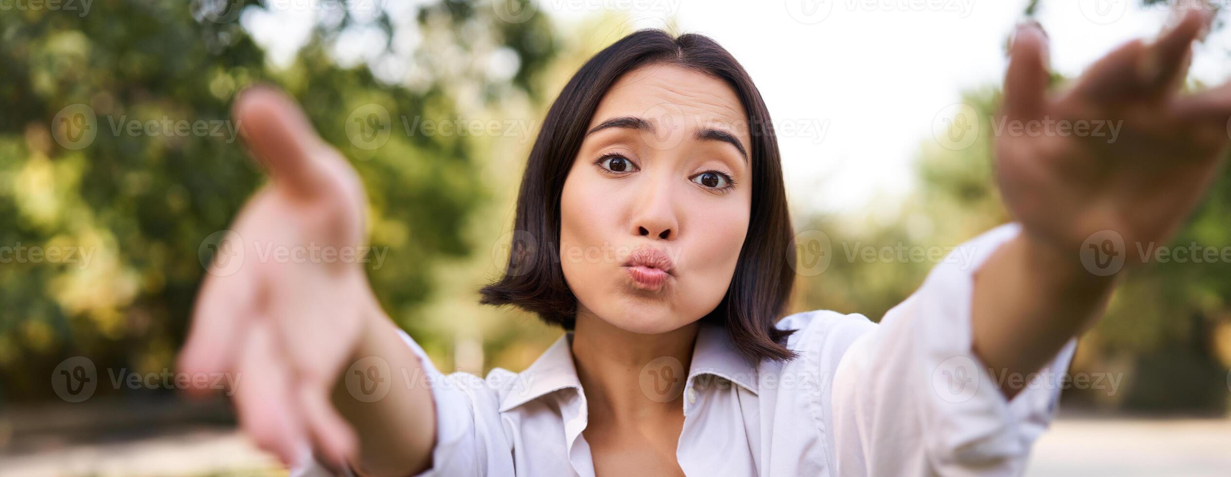 People and mobile connection. Happy young asian woman takes selfie on smartphone, holds camera with hands, poses in park on summer day photo