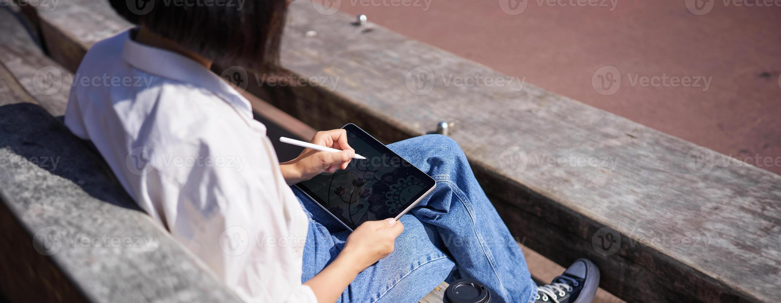 Cropped shot of female hands drawing on digital graphic tablet with pen, sitting with cup of coffee outdoors on bench photo