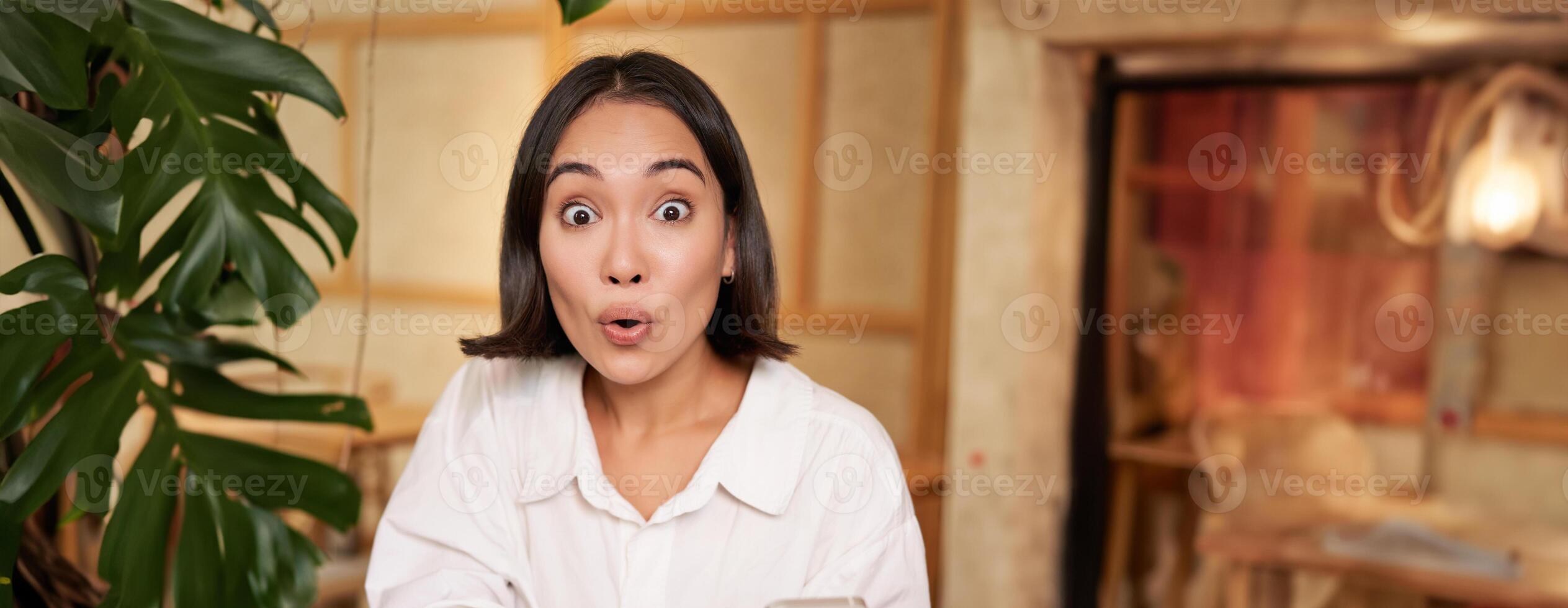 retrato de asiático niña mirando sorprendido, participación móvil teléfono con asombrado rostro, genial Noticias en teléfono inteligente, sentado en café con libro foto