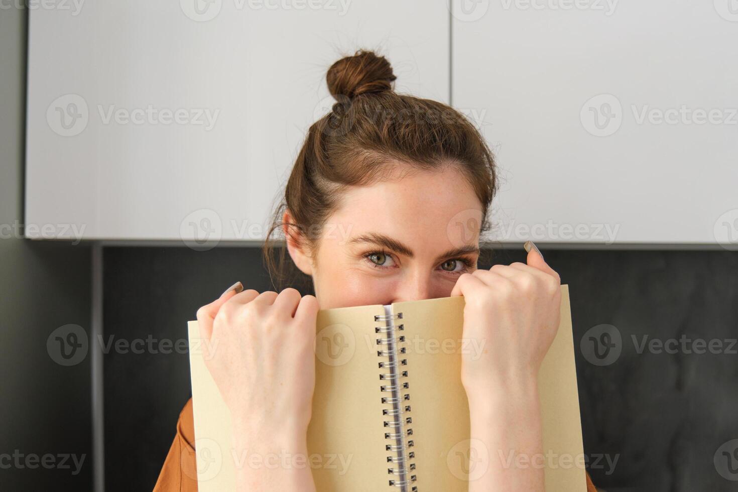 Cute romantic woman, hiding face behind notebook, smiling with eyes photo