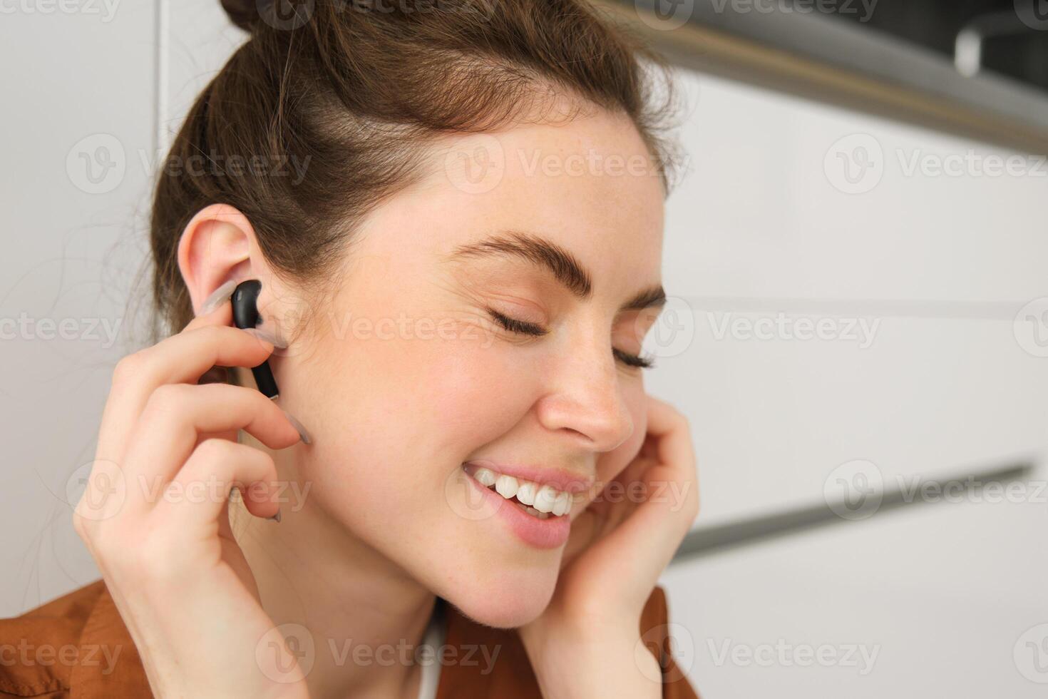 Portrait of beautiful woman enjoys listening to music or podcast in wireless black headphones, using earphones, smiling and looking happy, sitting at home on kitchen floor photo