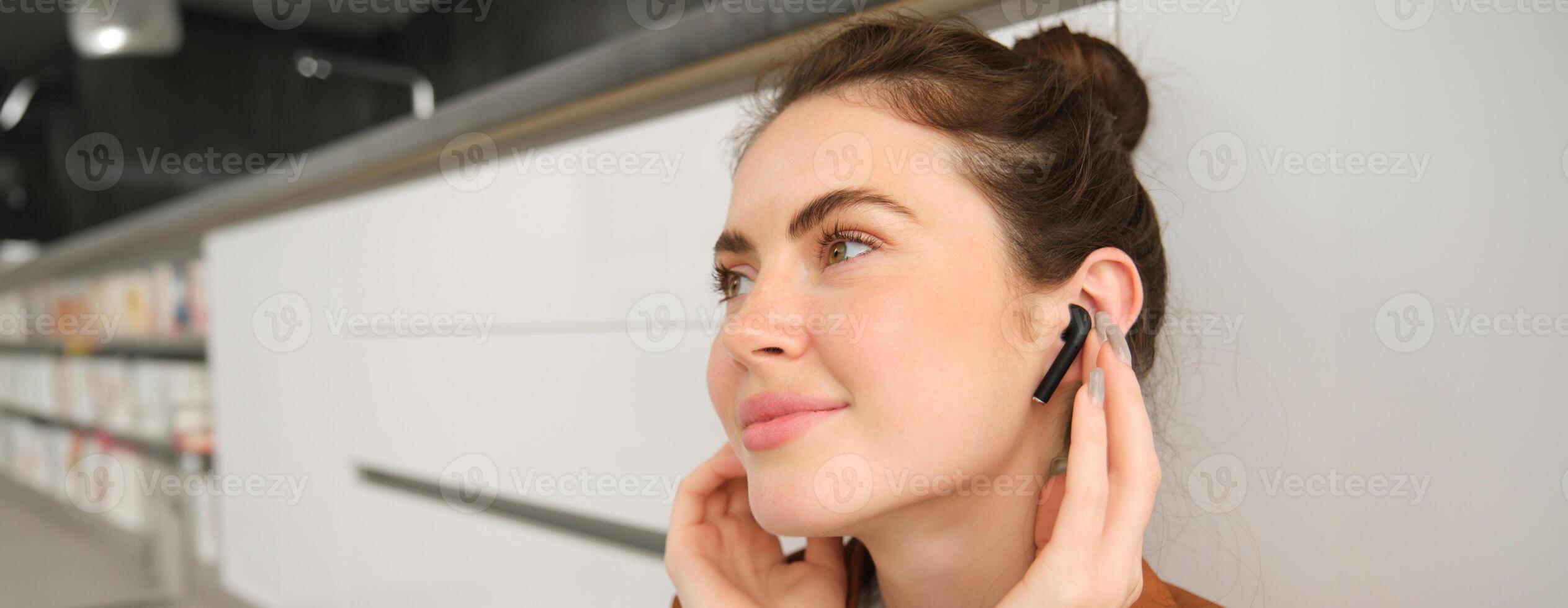 Close up photo of young smiling brunette woman, puts on her headphones, listens to music in wireless earphones, sits in the kitchen and enjoys sound quality