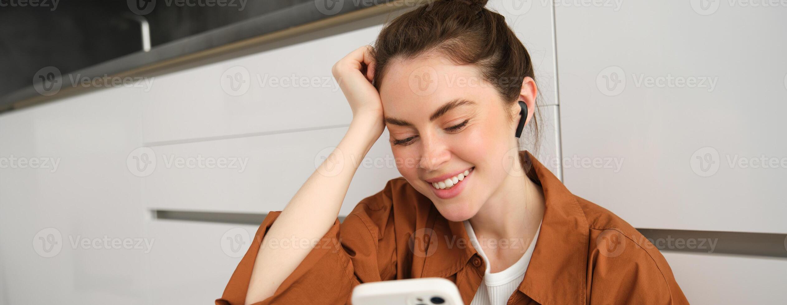 Portrait of happy young beautiful woman, listening music in wireless earphones, using smartphone, watching videos on mobile phone with headphones on photo