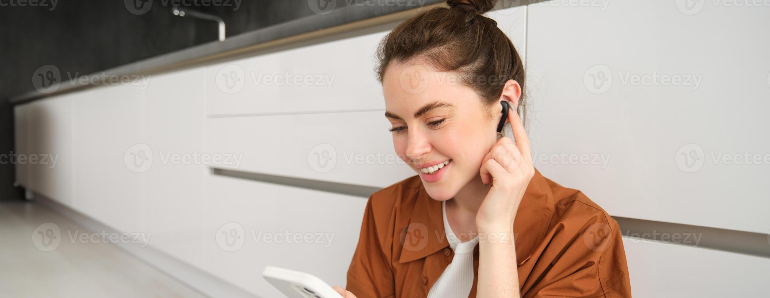 Portrait of woman enjoying watching videos on smartphone with wireless earphones, girl listening to music and using mobile phone, sitting on kitchen floor photo