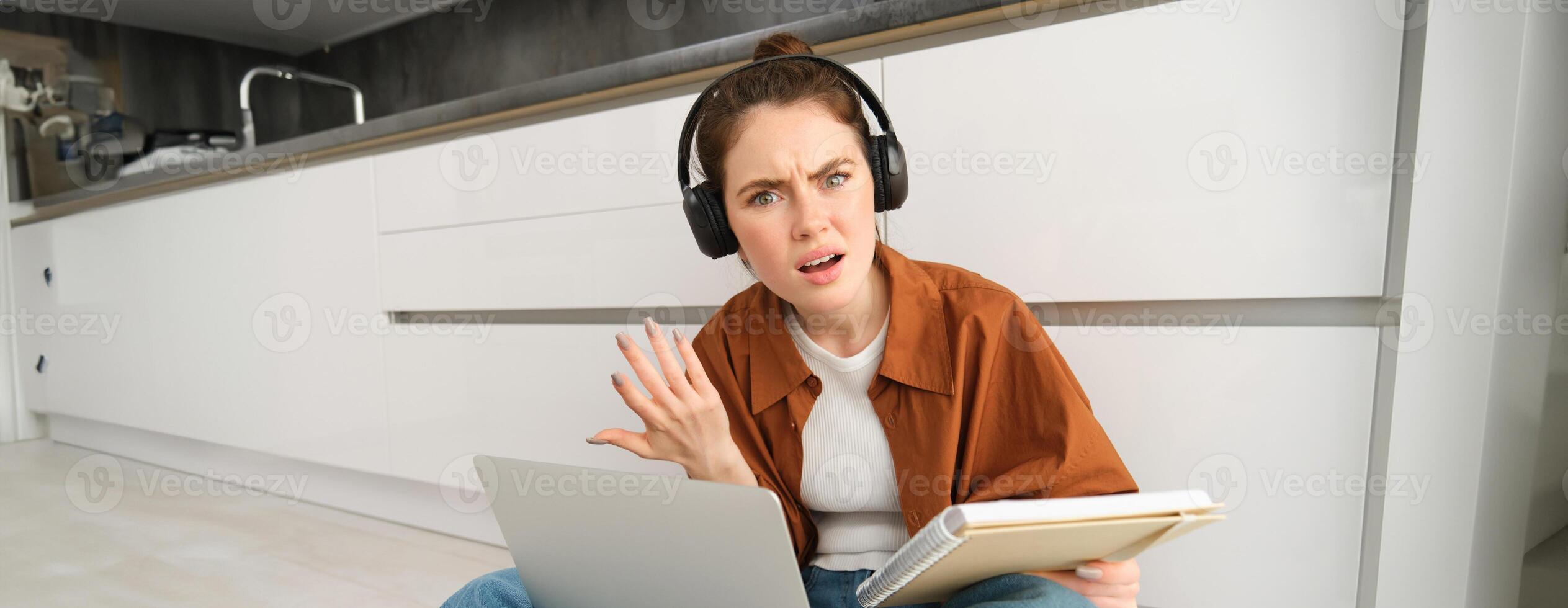 Portrait of annoyed and confused woman in headphones, sits on floor with laptop and notebook, looks frustrated, has issues in doing homework or studying photo