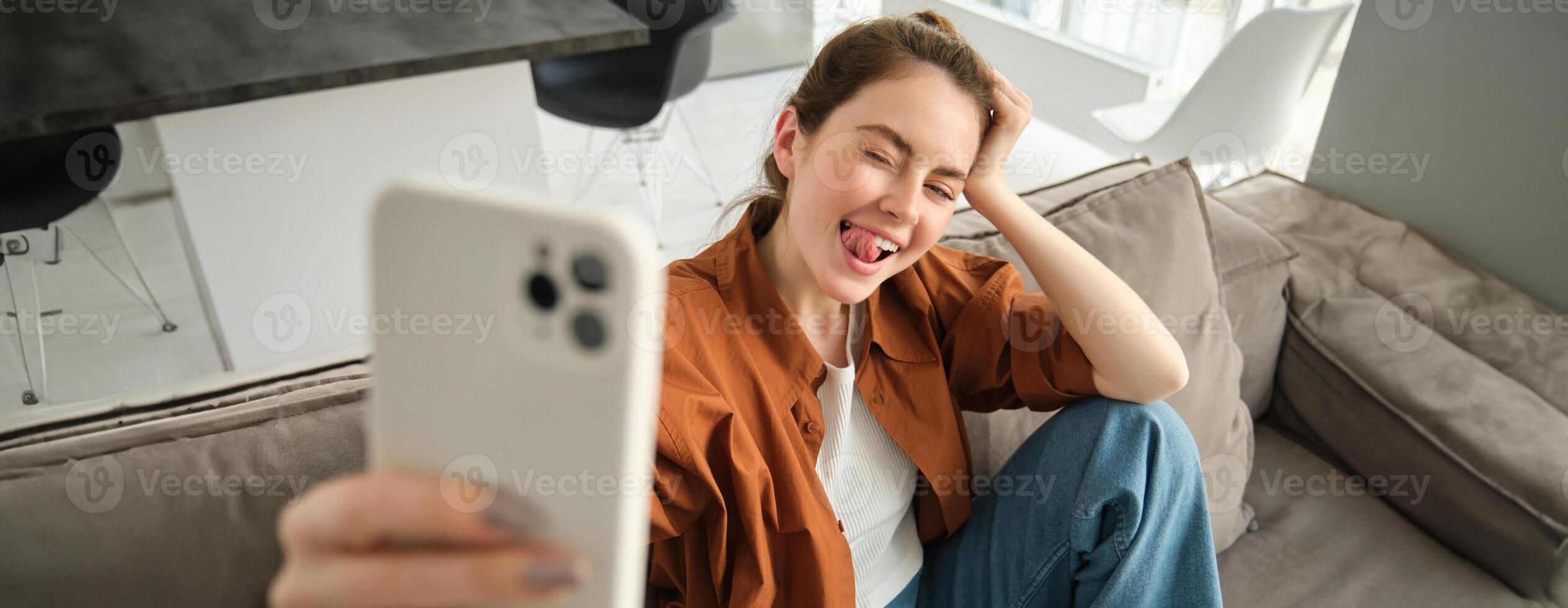 despreocupado moderno mujer, sonriente y tomando selfies en teléfono inteligente aplicación, posando para foto en su sofá, sentado en sofá con móvil teléfono