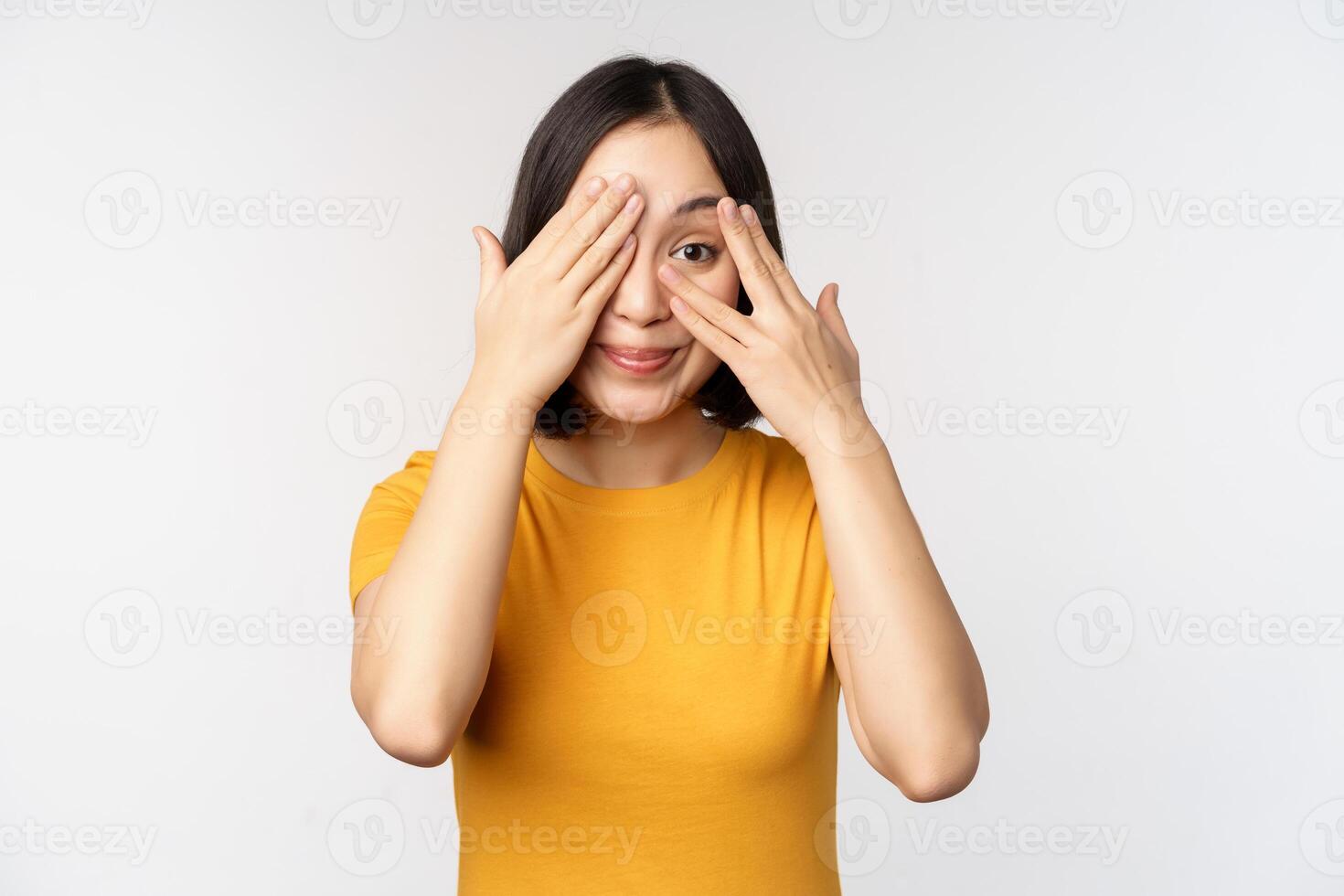 Cute korean female model, cover eyes with hands, peeking through fingers curious, watching smth, standing in yellow tshirt over white background photo