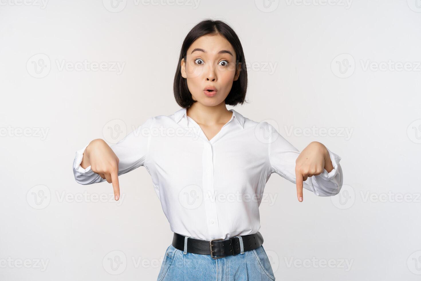 Portrait of young asian woman pointing fingers down and smiling, showing banner, click on link below gesture, inviting people to follow, standing over white background photo