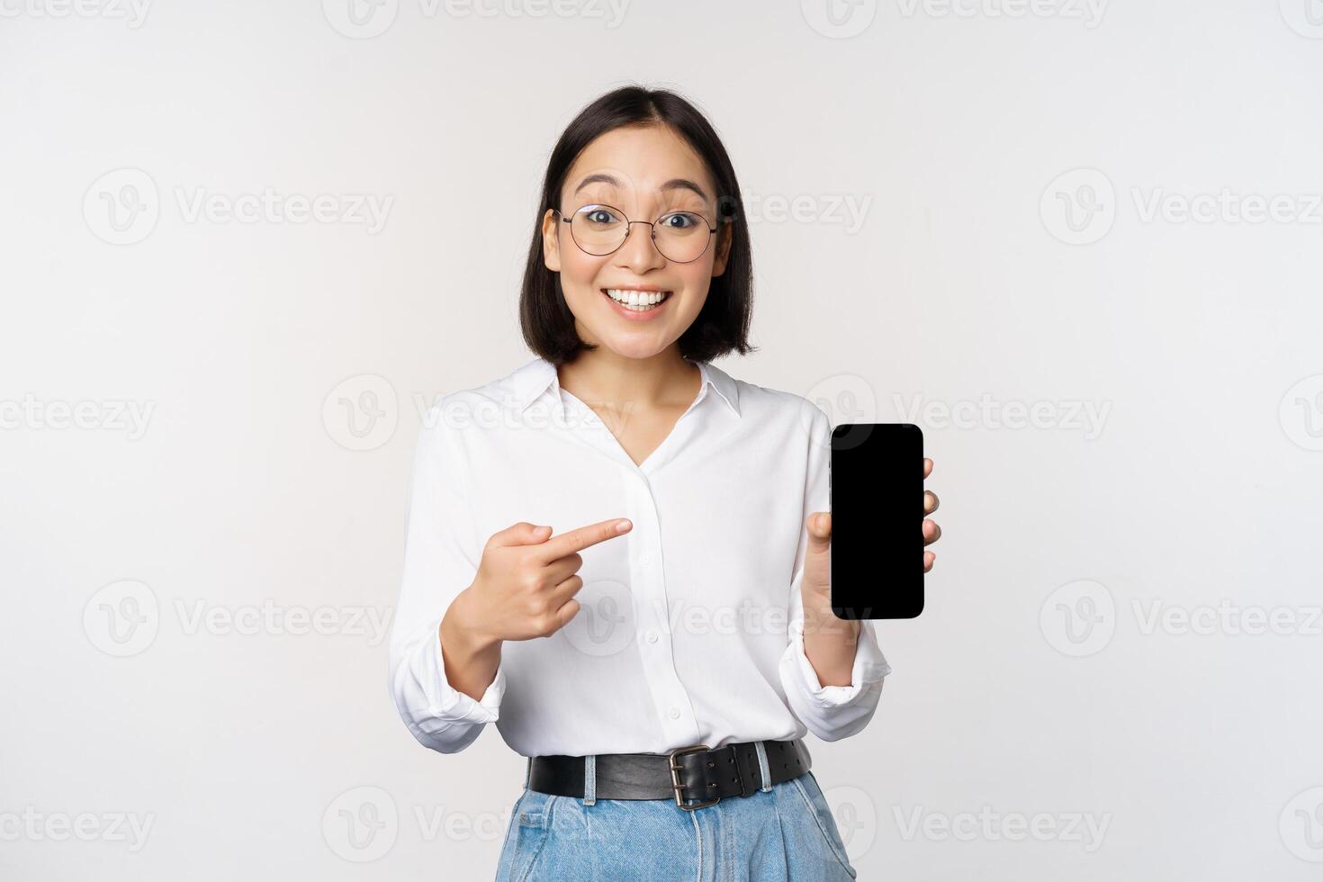 Enthusiastic young asian woman pointing finger at smartphone screen, showing advertisement on mobile phone, white background photo