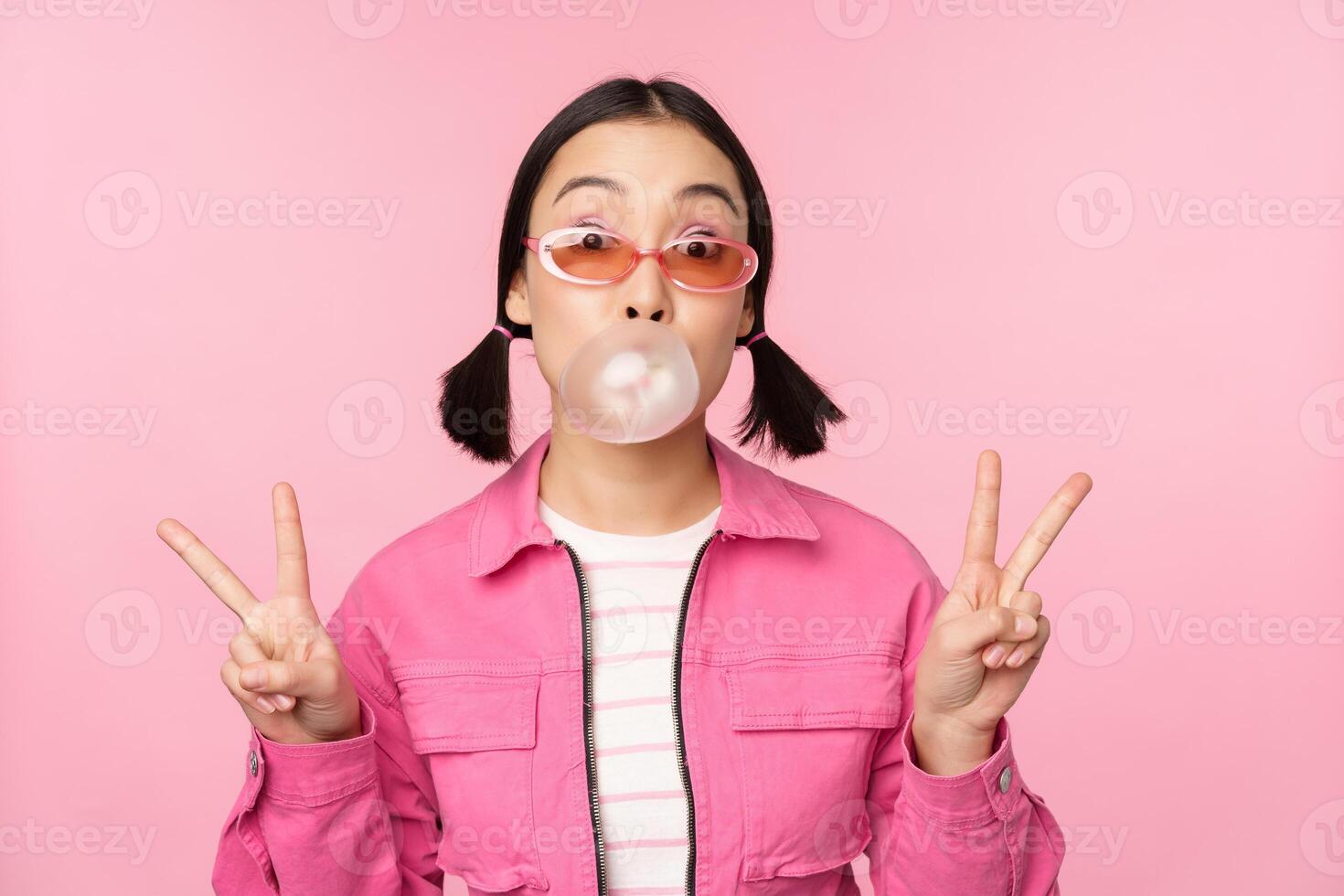 hermosa coreano niña en Gafas de sol, soplo chicle burbuja y demostración paz señales, en pie terminado rosado antecedentes foto