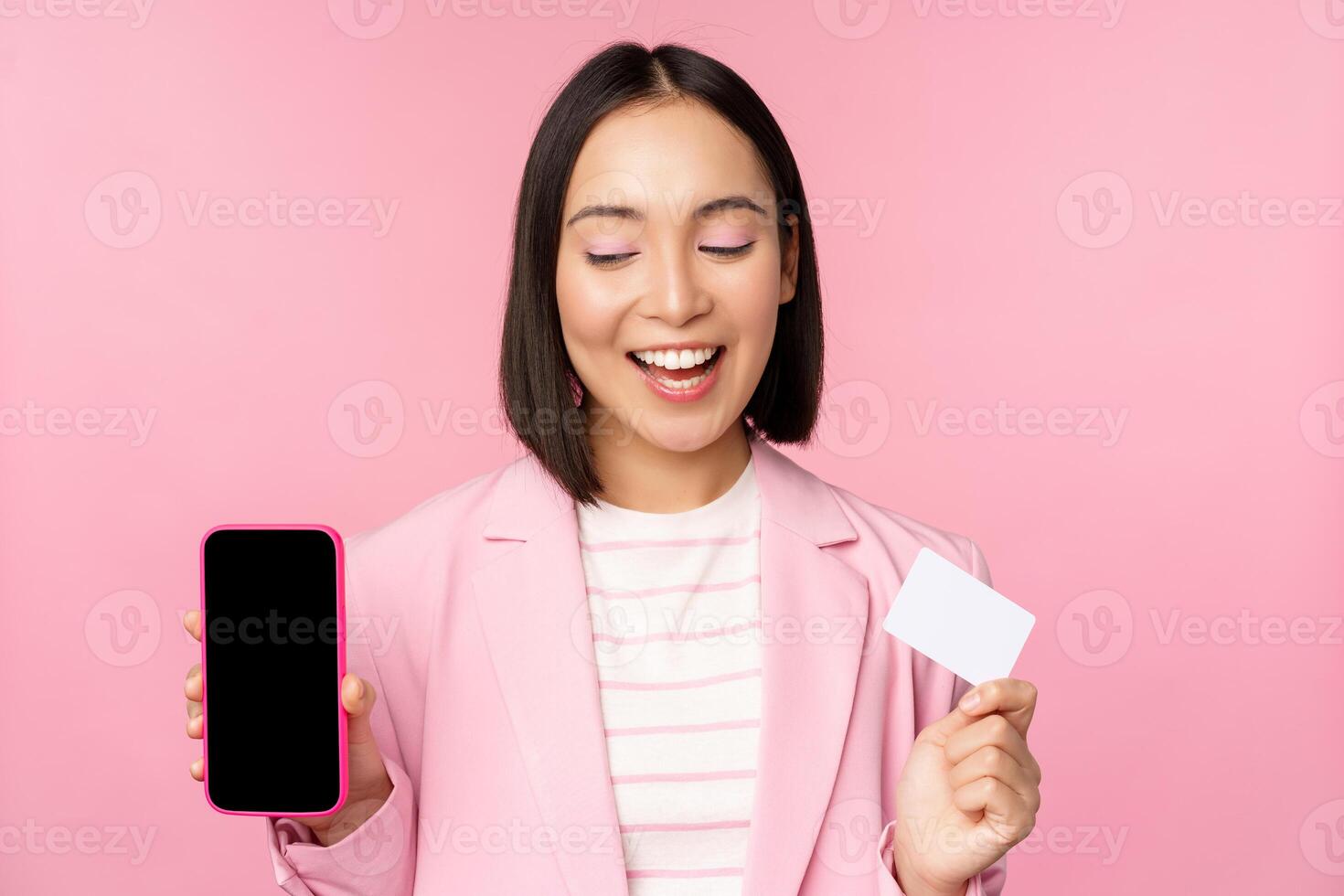 Smiling korean businesswoman in suit, showing mobile phone screen, credit card, showing online banking application interface, pink background photo