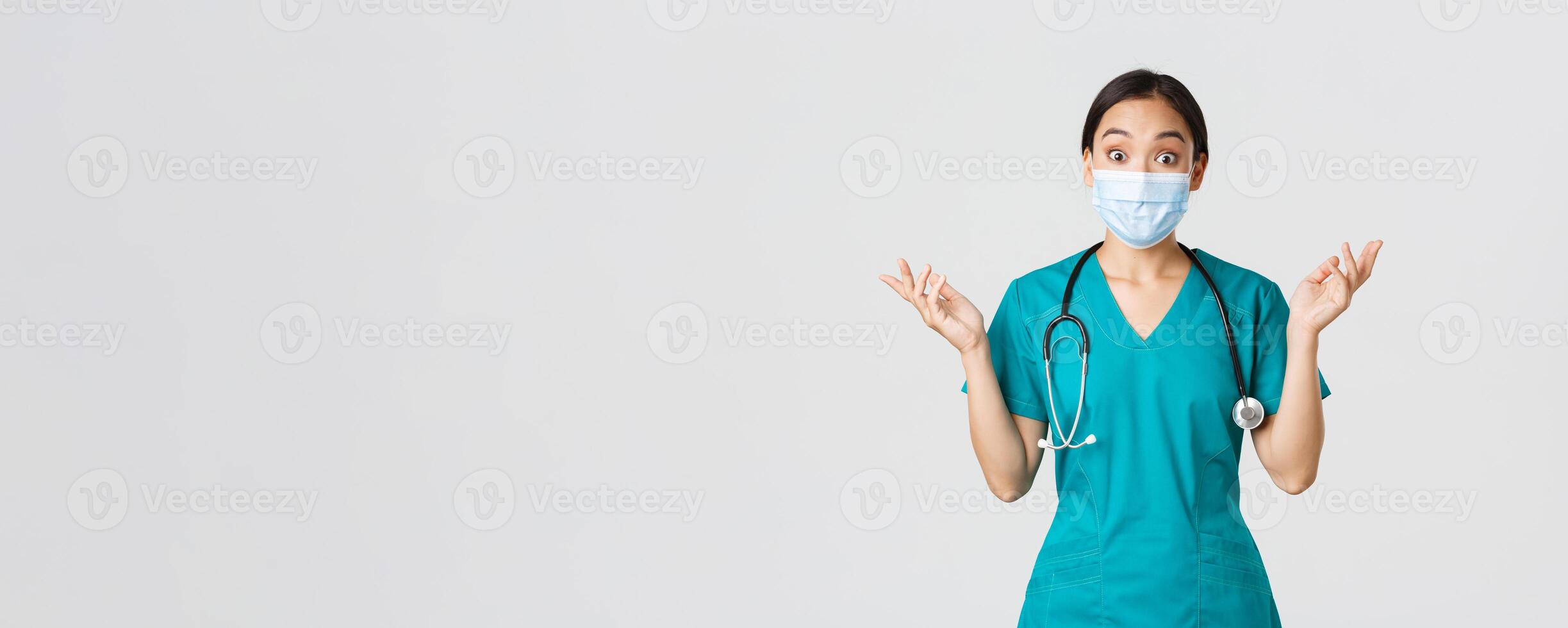 Covid-19, coronavirus disease, healthcare workers concept. Surprised and amazed asian female nurse, physician in medical mask and scrubs raising hands up excited, hear amazing news, white background photo