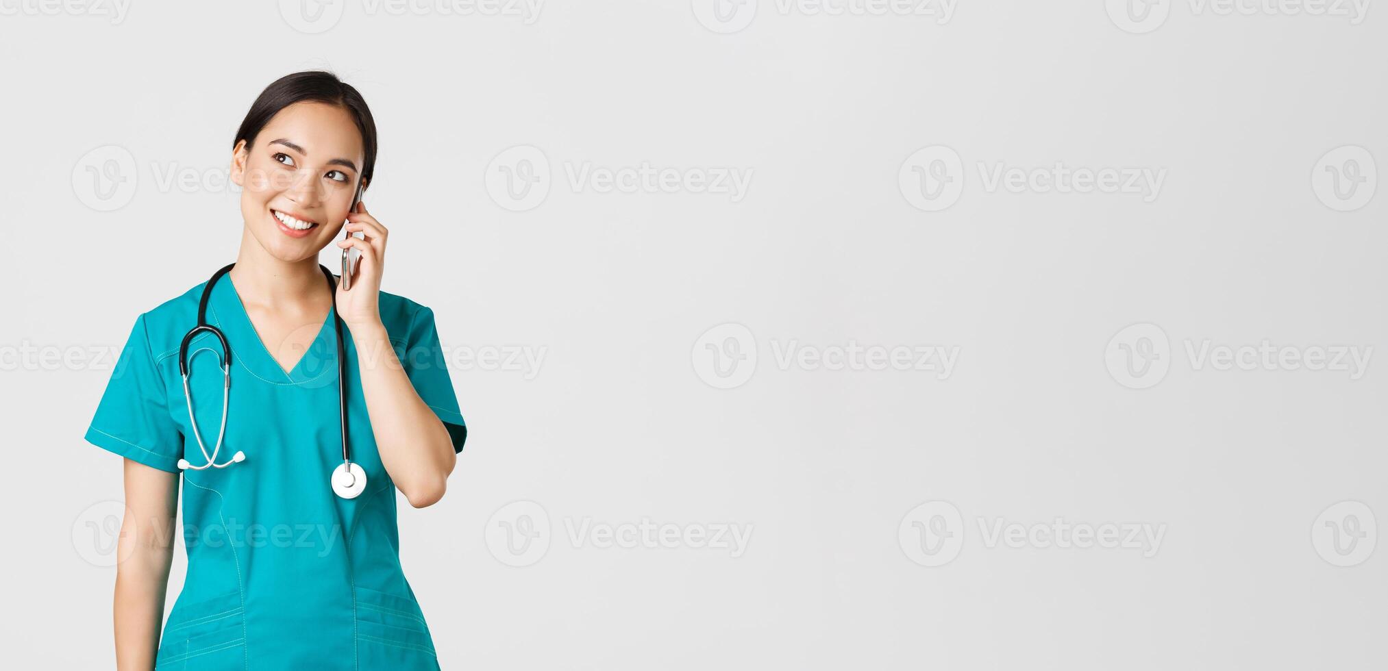 Covid-19, healthcare workers and preventing virus concept. Pretty smiling asian female doctor, physician in scrubs having conversation, talking on phone and looking upper left corner dreamy photo
