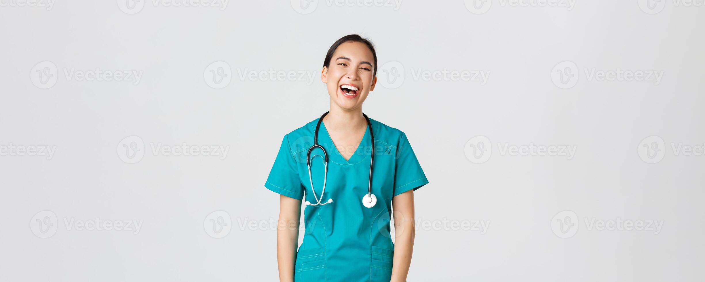 Covid-19, healthcare workers, pandemic concept. Carefree beautiful female asian nurse in scrubs, intern having fun, laughing happy. Doctor on lunch break chuckle and talking to coworkers photo