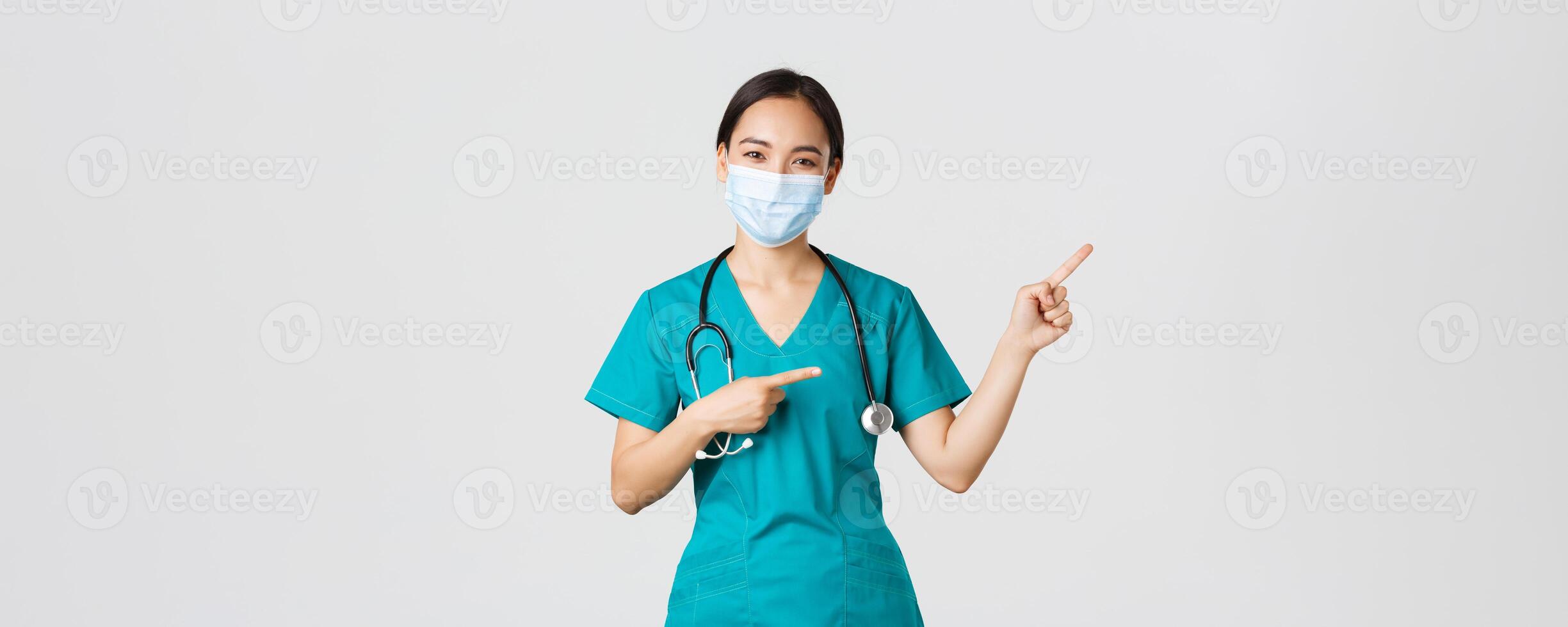 Covid-19, coronavirus disease, healthcare workers concept. Pleasant asian female doctor, medical employee in scrubs and mask, pointing fingers upper right corner, showing advertisement photo