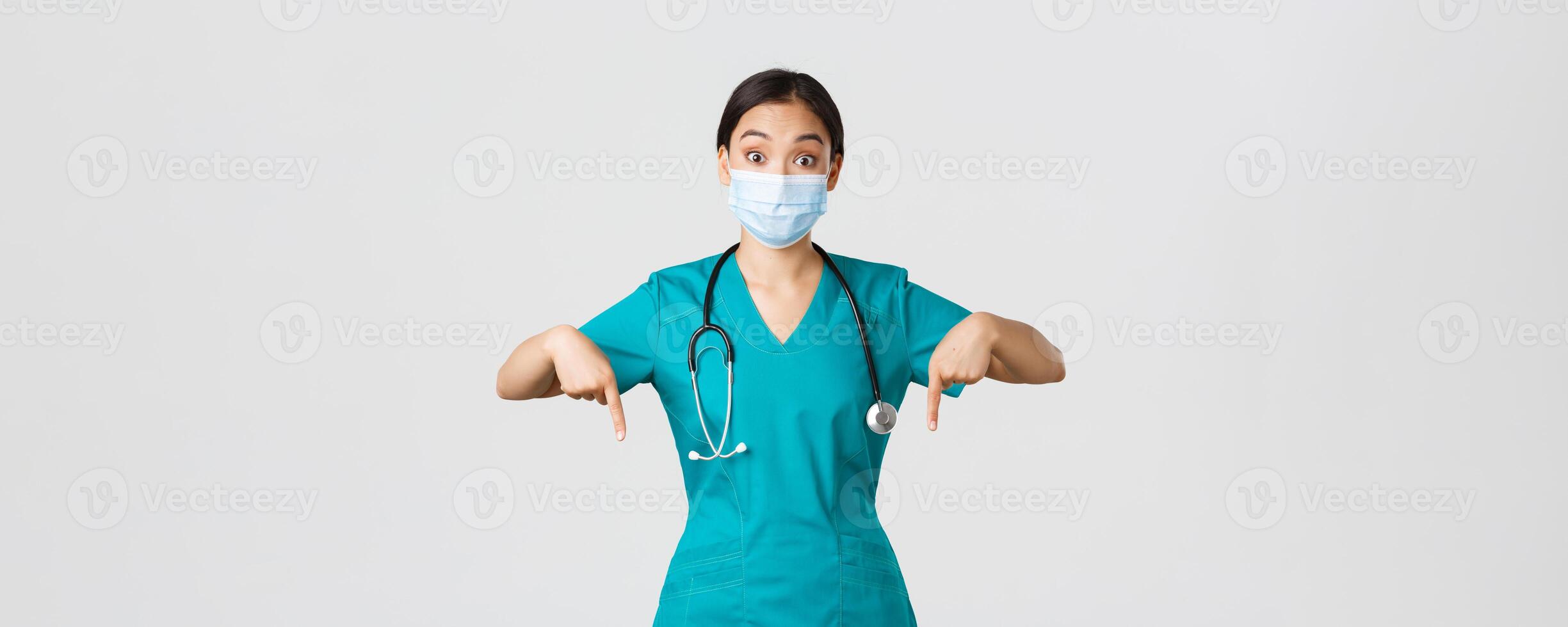 Covid-19, coronavirus disease, healthcare workers concept. Excited and surprised asian female physician, nurse in scrubs and medical mask, pointing fingers down, showing advertisement photo