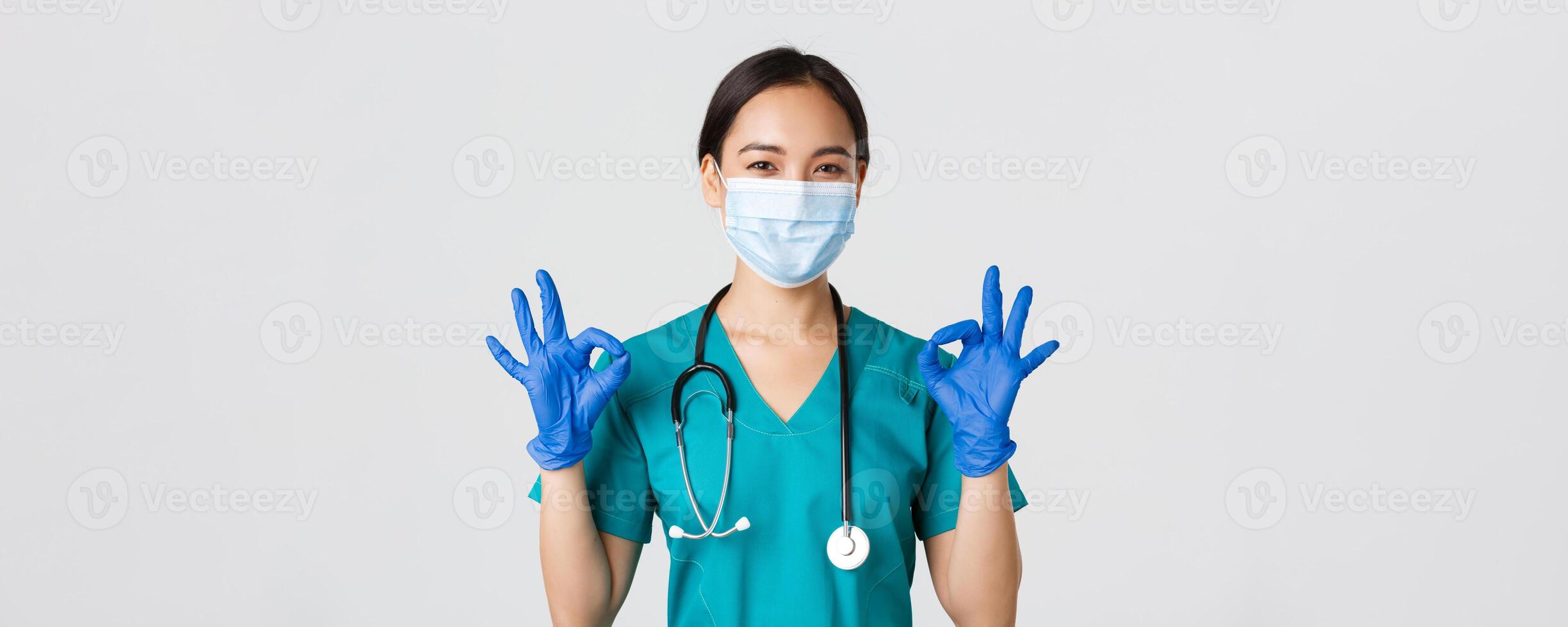Covid-19, coronavirus disease, healthcare workers concept. Confident smiling asian female doctor, nurse in medical mask and gloves, show okay gesture in approval, white background photo