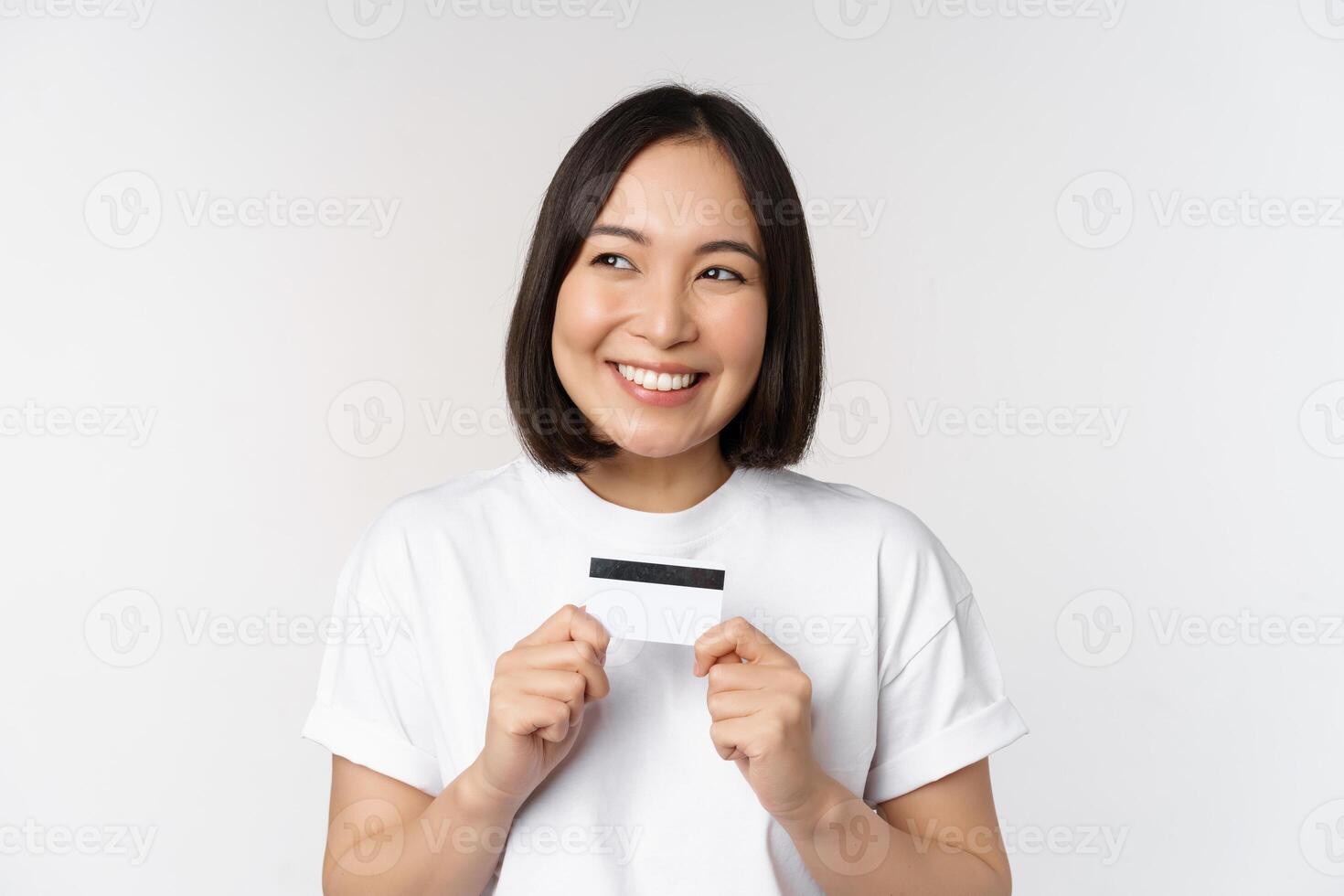 sonriente coreano mujer demostración crédito tarjeta con contento rostro, en pie en camiseta terminado blanco antecedentes foto