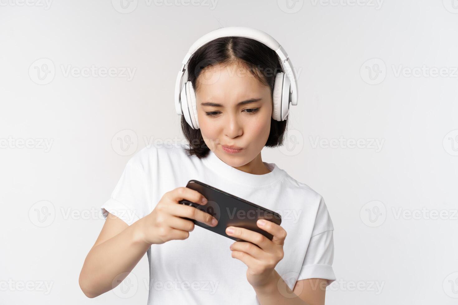 contento asiático mujer en auriculares, mirando a teléfono inteligente, acecho vídeo en móvil teléfono y sonriente, en pie terminado blanco antecedentes foto