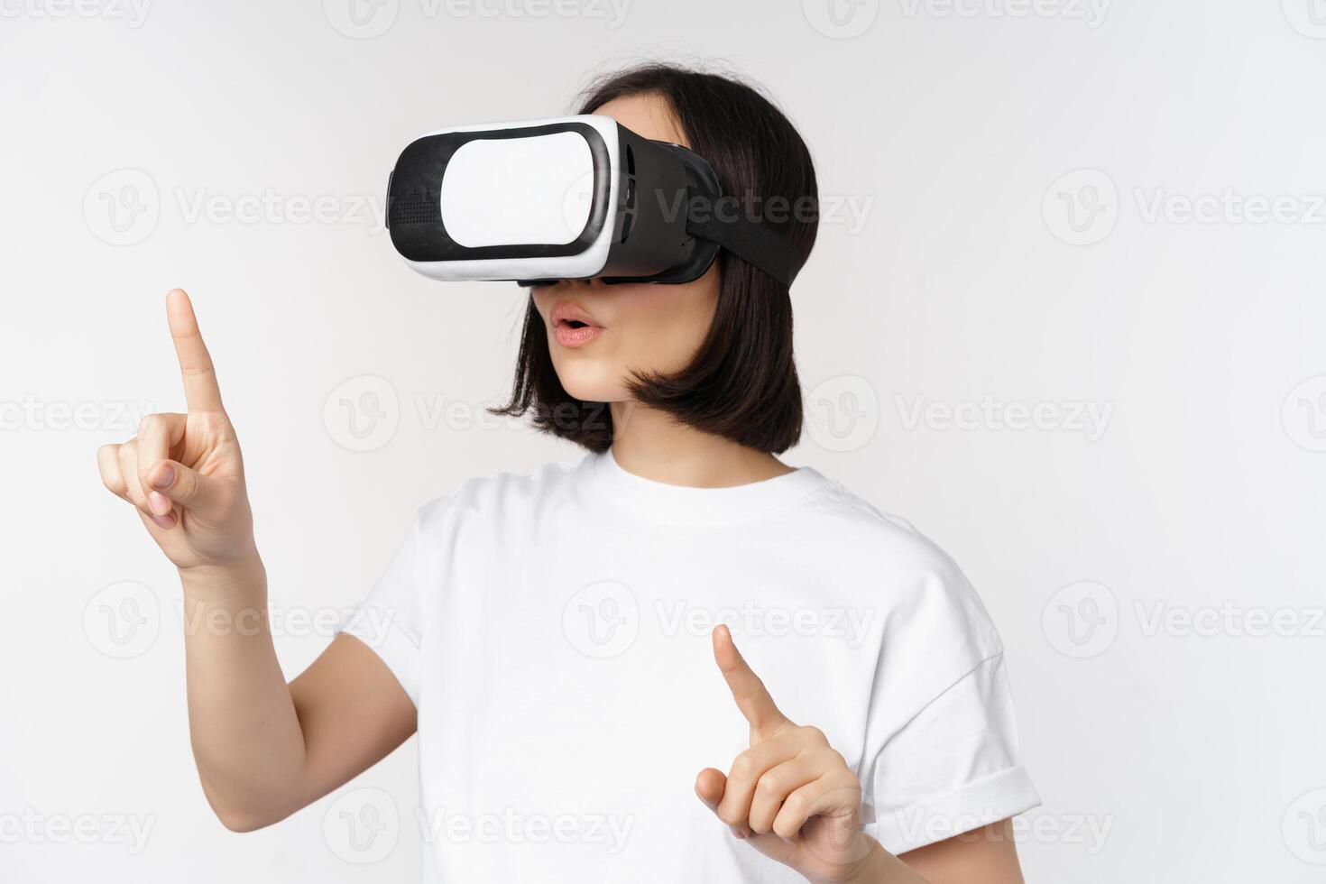 Potrait of asian woman in virtual reality glasses, pointing, choosing smth in VR headset, standing over white background photo