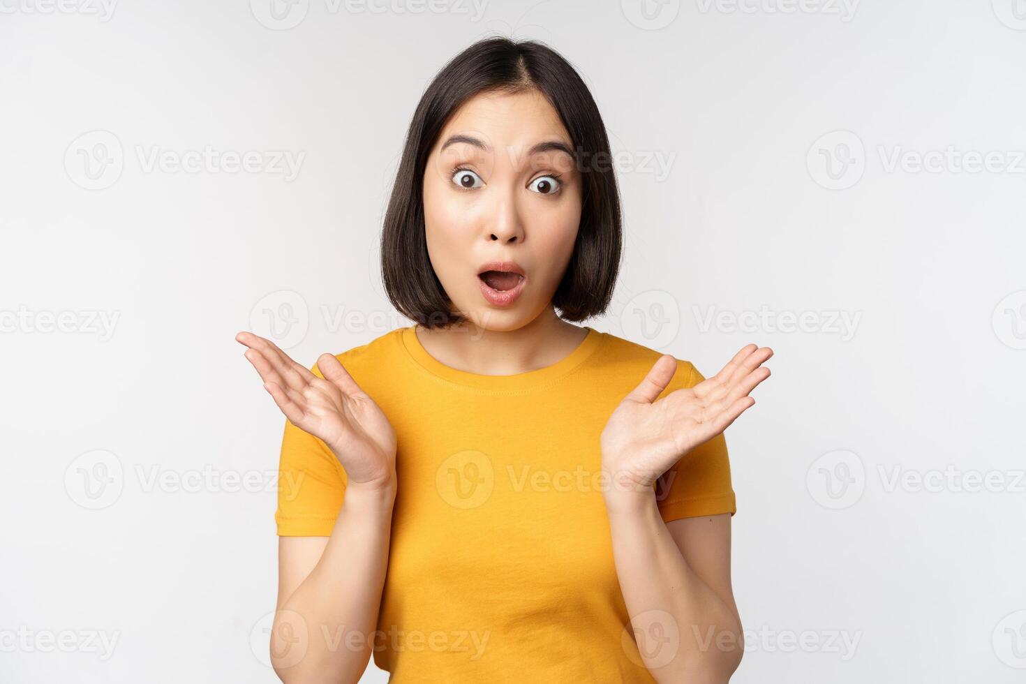 Close up portrait of asian woman looking surprised, wow face, staring impressed at camera, standing over white background in yellow t-shirt photo