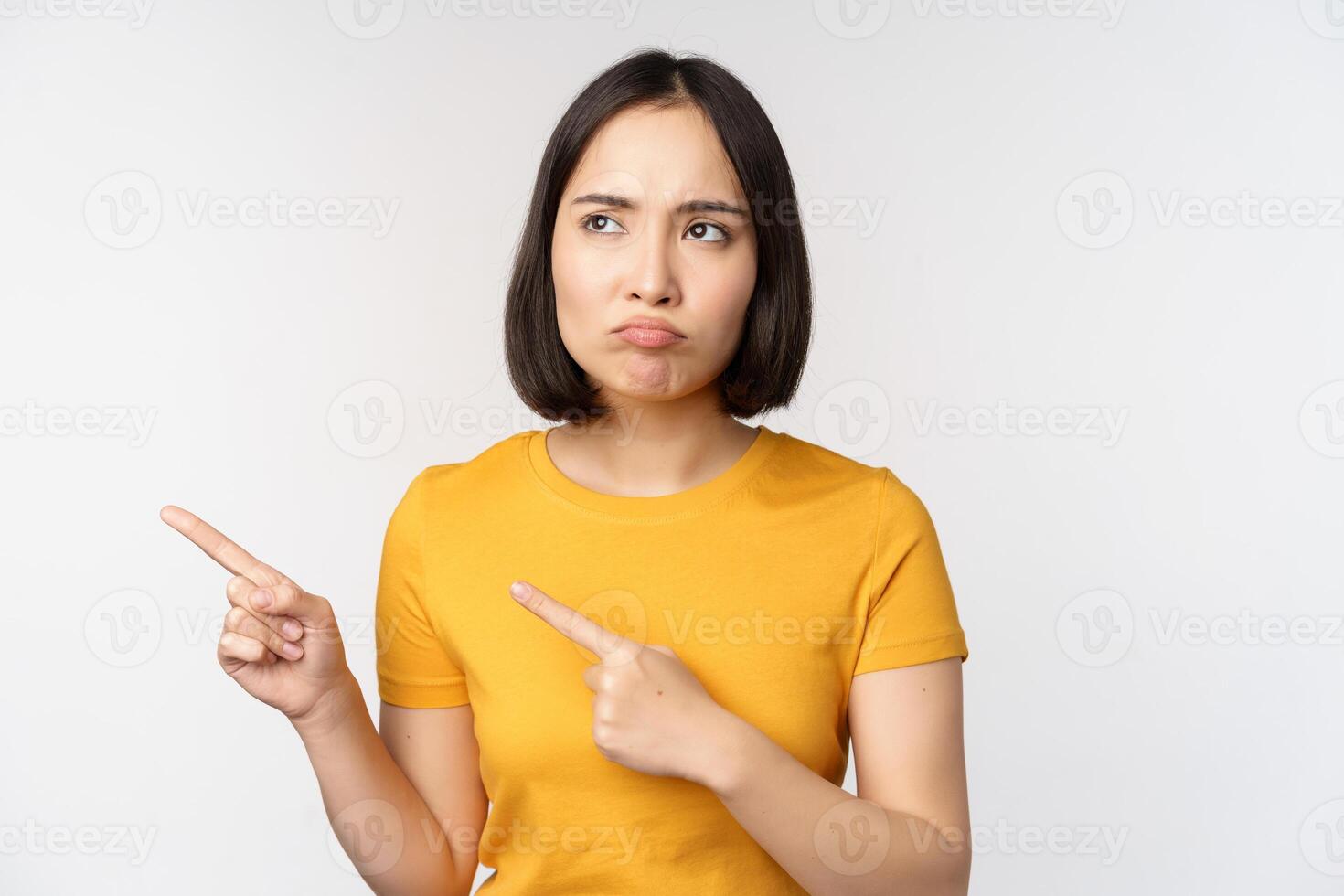 Upset grumpy asian girl, pouting and looking at smth unfair, pointing fingers left at banner, logo brand, standing in yellow tshirt over white background photo