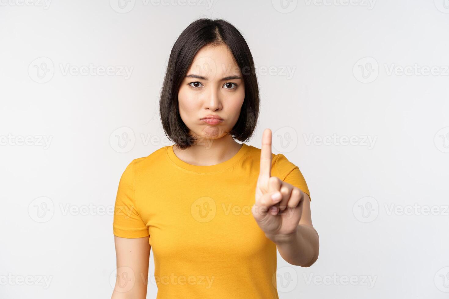 Portrait of asian woman looking serious and angry, showing stop prohibit gesture, taboo sign, forbidding smth, standing in yellow tshirt over white background photo