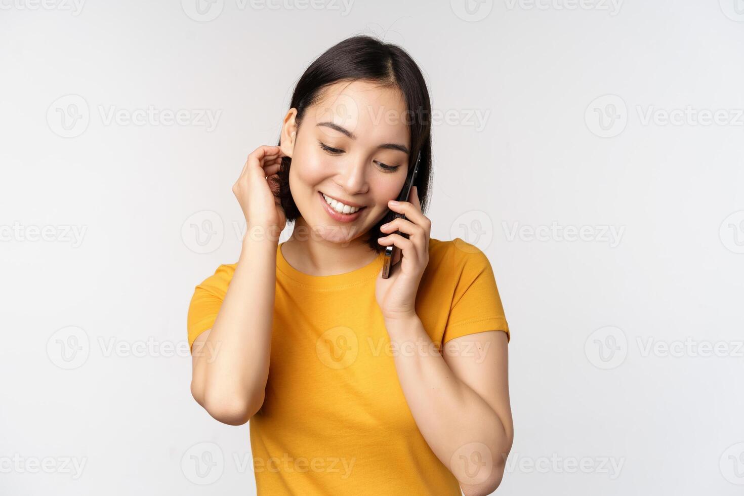 Romantic and cute asian girl answer phone call, talking on mobile, smiling coquettish, standing in yellow tshirt against white background photo