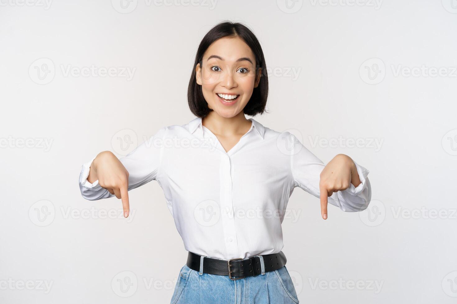 Portrait of young asian woman pointing fingers down and smiling, showing banner, click on link below gesture, inviting people to follow, standing over white background photo
