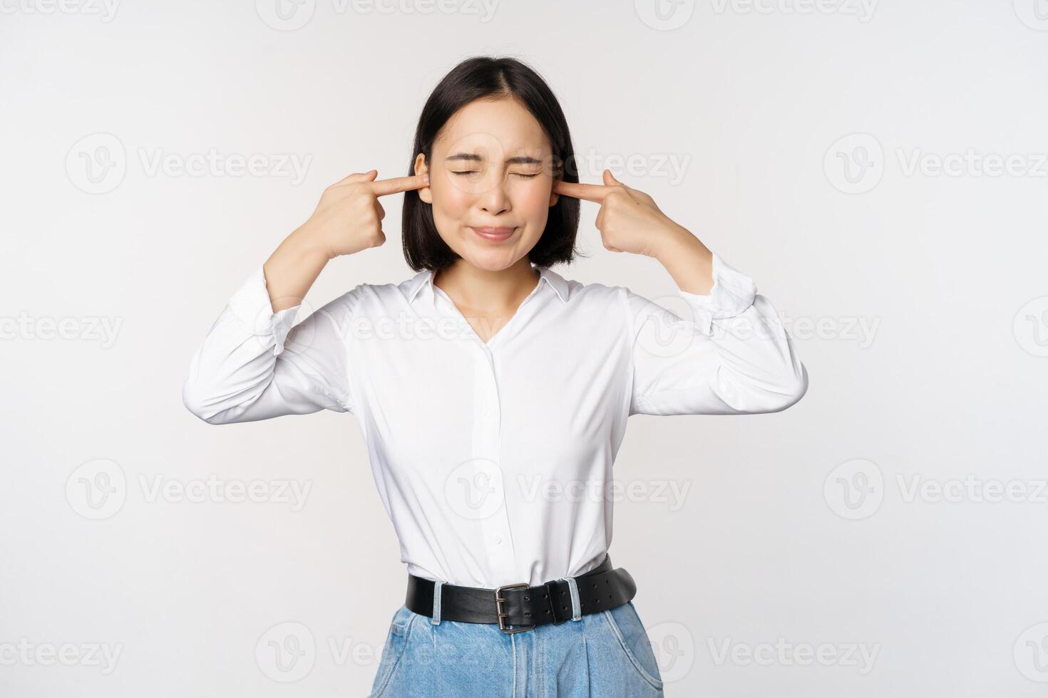 Annoyed asian woman feeling discomfort from loud annoying noise, close ears and eyes, standing over white background photo