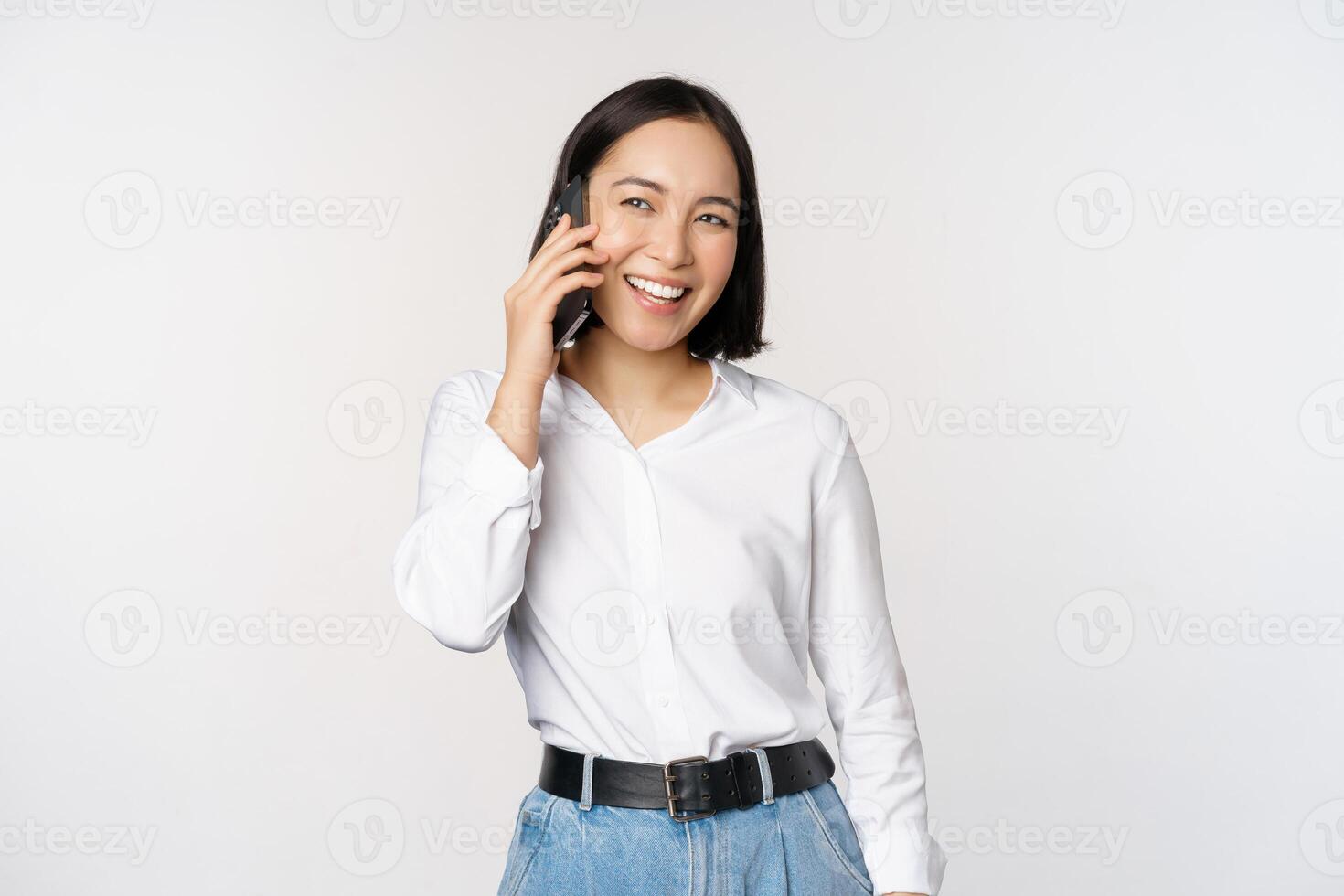 Friendly smiling asian woman talking on phone, girl on call, holding smartphone and laughing, speaking, standing over white background photo