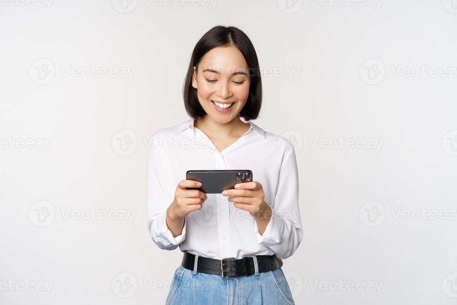 Young korean woman, asian girl playing mobile video game on smartphone, looking at horizontal phone screen, standing over white background photo
