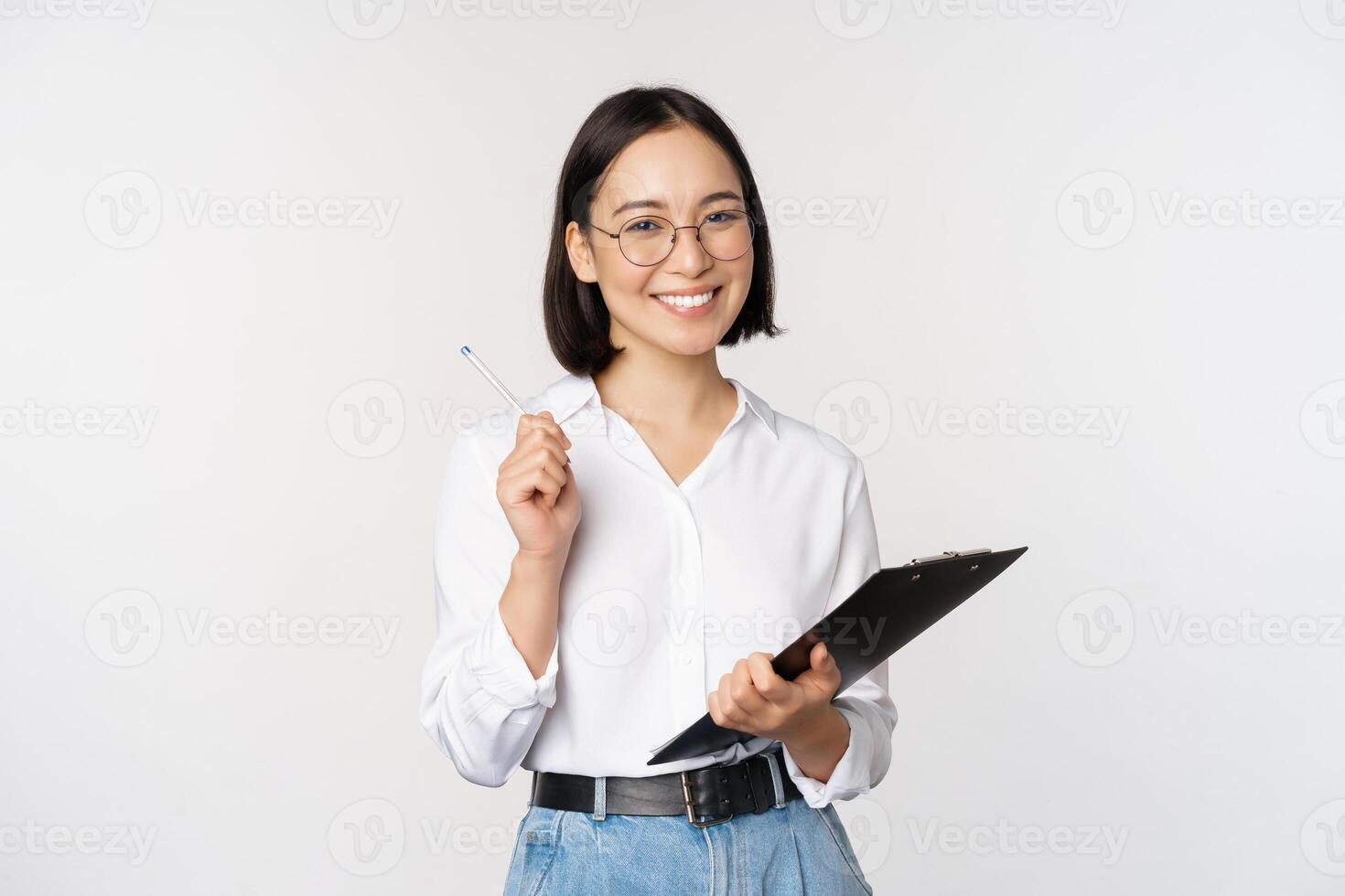 Smiling young asian woman taking notes with pen on clipboard, looking happy, standing against white background photo