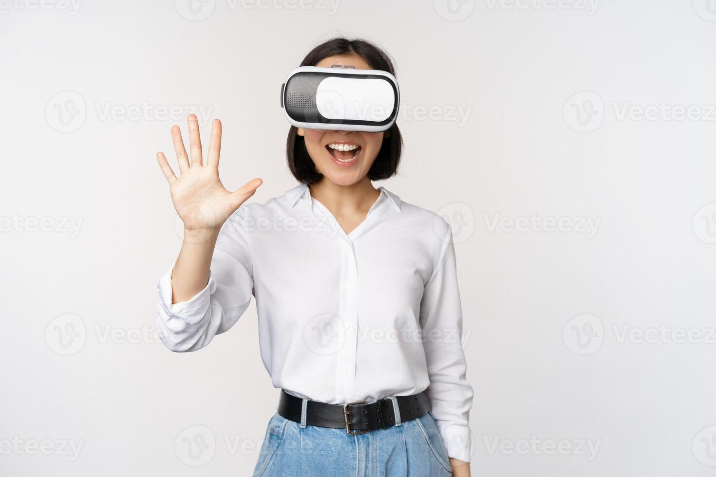 Smiling asian woman waving at you, wearing virtual reality glasses, vr headset, chatting in meta universe, standing over white background photo