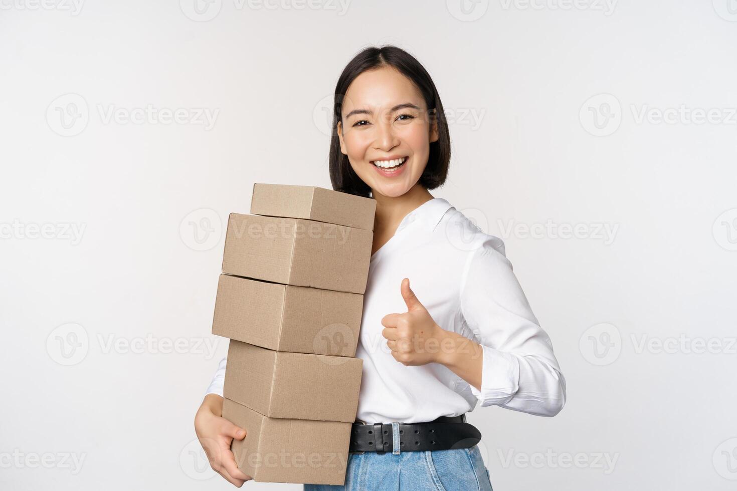 imagen de contento moderno asiático mujer demostración pulgares arriba, participación cajas entrega bienes, en pie en contra blanco antecedentes foto