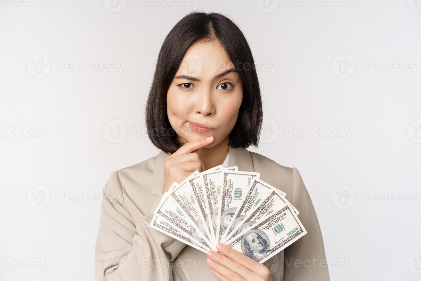 Image of asian corporate woman, happy businesswoman showing money, cash dollars and thinking, standing in suit over white background photo