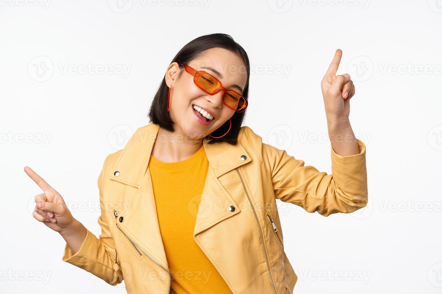 sonriente asiático morena mujer en Gafas de sol, señalando dedos oblicuo, izquierda y bien, demostración variantes, riendo y baile, vistiendo Gafas de sol, blanco antecedentes foto