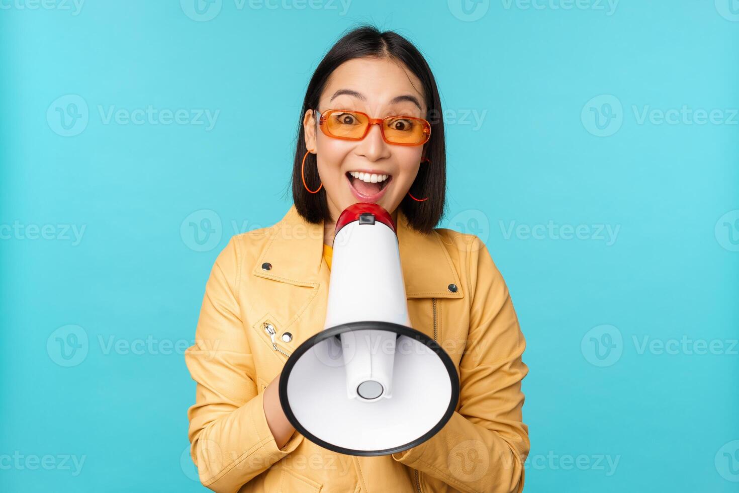 elegante asiático niña haciendo anuncio en megáfono, gritos con altavoz y sonriente, atractivo gente, reclutamiento, en pie terminado azul antecedentes foto
