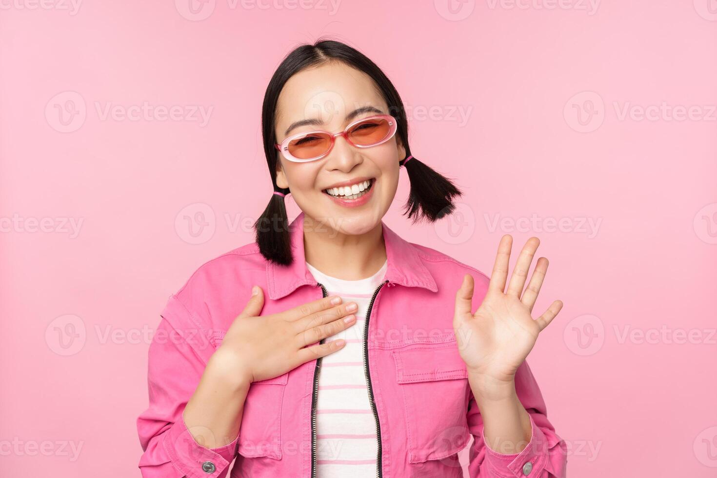 Portrait of friendly asian teen girl in sunglasses saying hello, waves her hand and smiles, greets you, hi gesture, stands over pink background photo