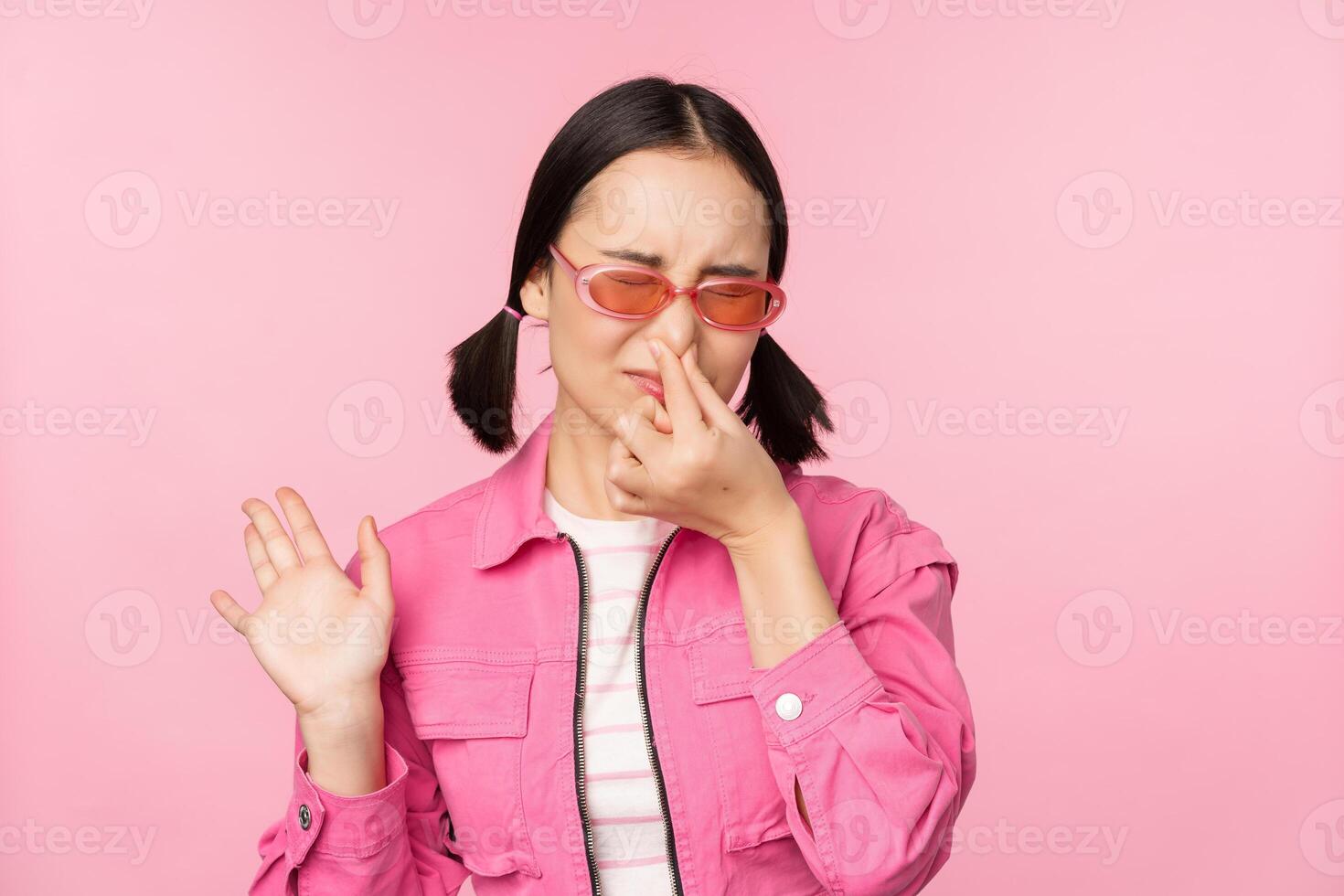 apestoso. asiático niña en Gafas de sol y elegante atuendo, cierra su nariz desde asco, disgusto malo oler, en pie terminado rosado antecedentes foto