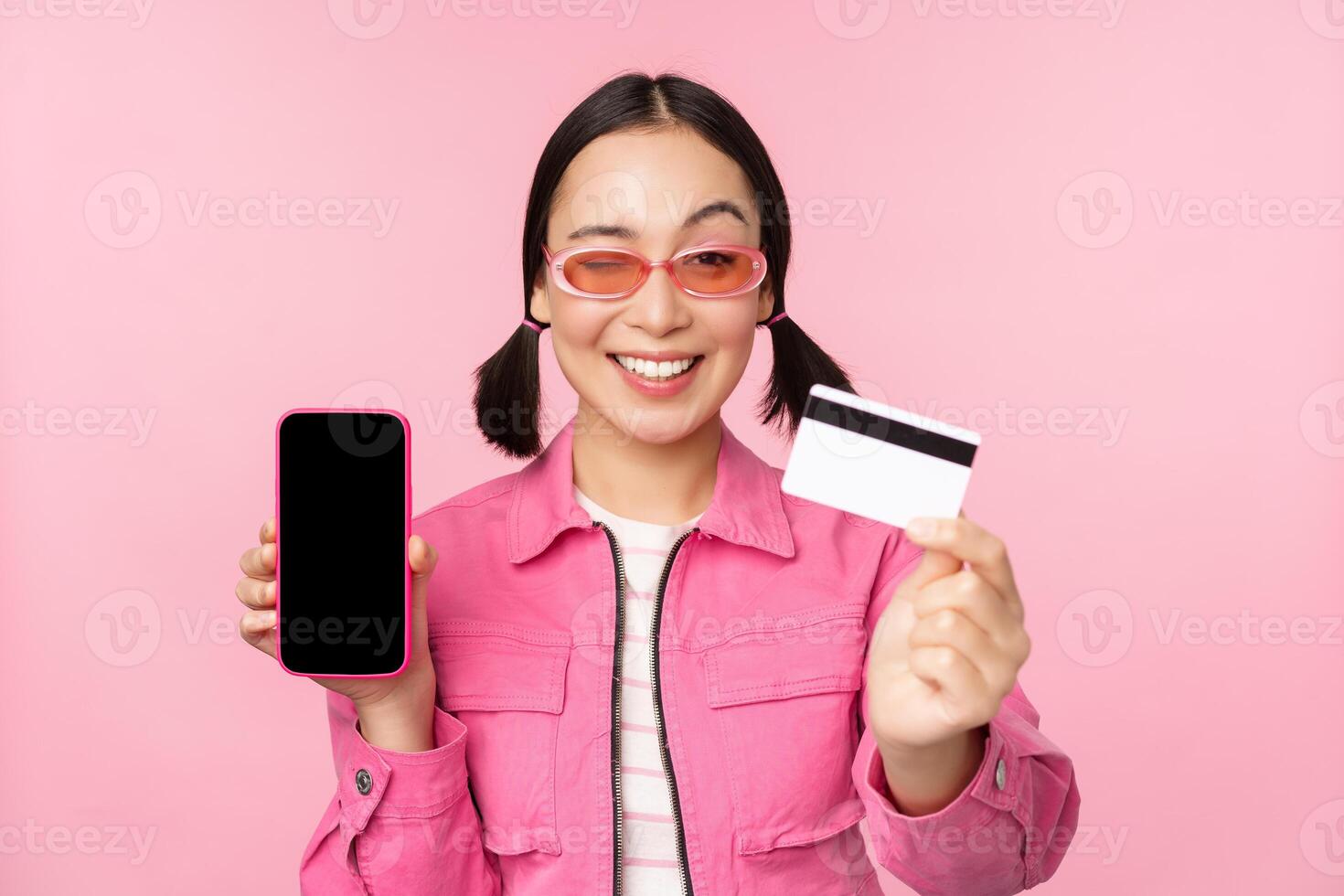 Image of smiling korean woman showing credit card and mobile phone screen, smartphone application interface, paying online, shopping contactless, standing over pink background photo