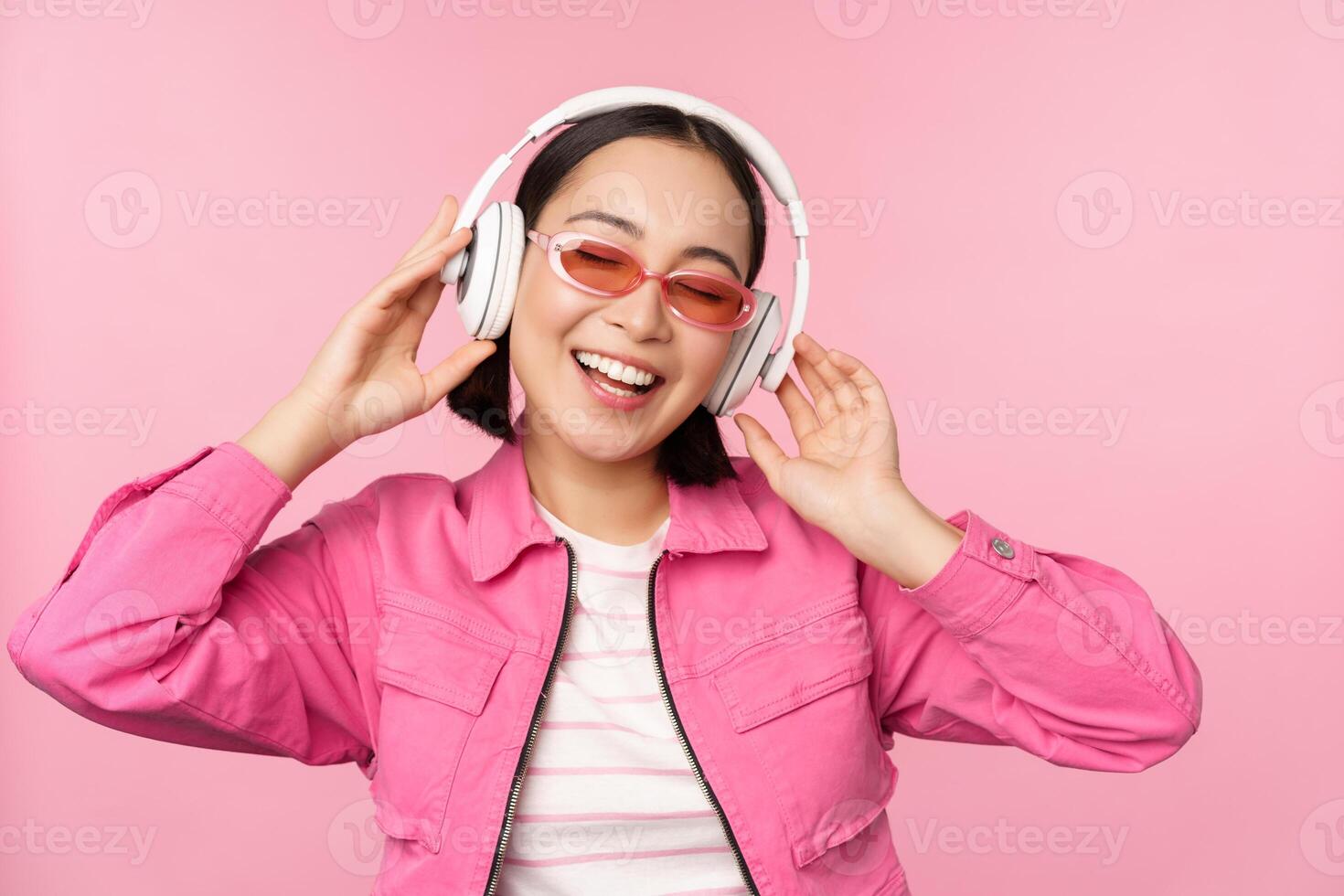 bailando elegante asiático niña escuchando música en auriculares, posando en contra rosado antecedentes foto