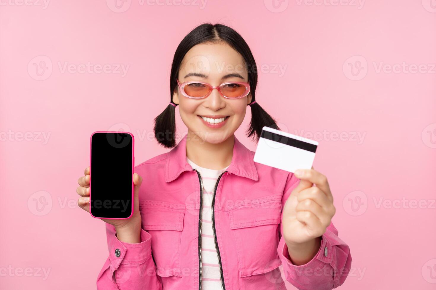 Image of smiling korean woman showing credit card and mobile phone screen, smartphone application interface, paying online, shopping contactless, standing over pink background photo