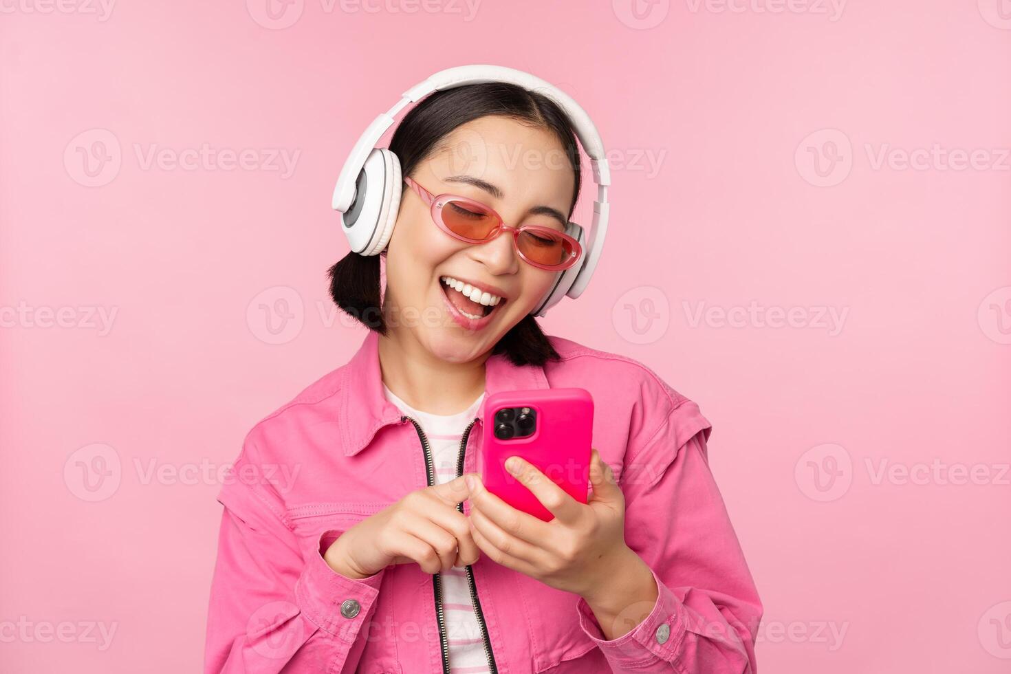 elegante asiático niña bailando con teléfono inteligente, escuchando música en auriculares en móvil teléfono aplicación, sonriente y reír, posando en contra rosado antecedentes foto