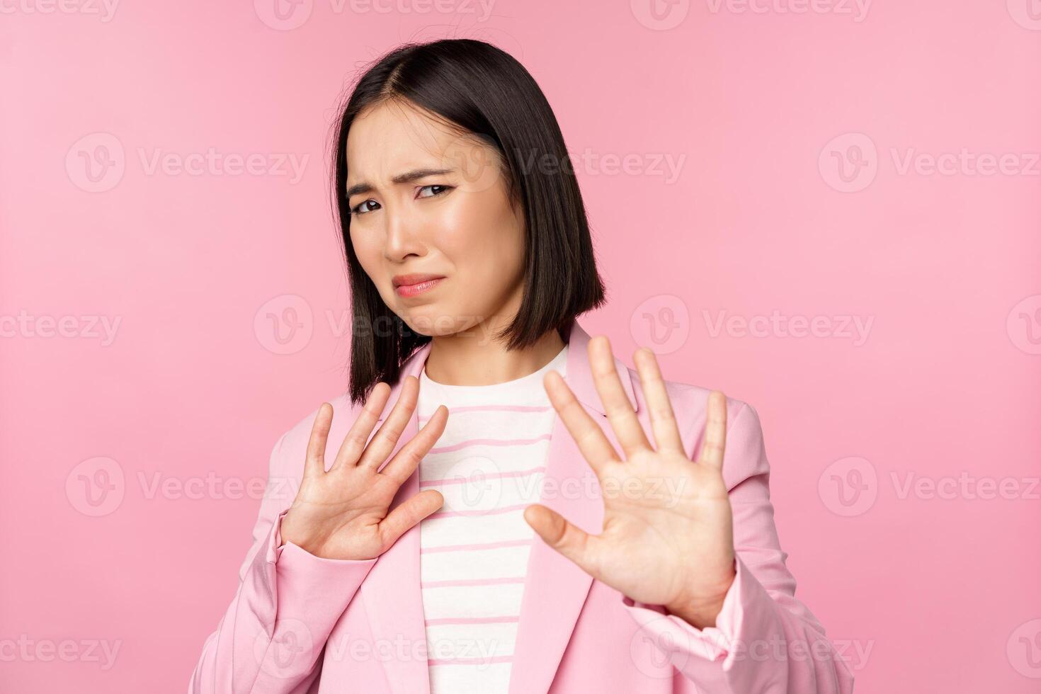 Get away. Disgusted asian businesswoman rejecting, shaking hands to refuse, grimacing from dislike, decline smth with aversion, posing in suit against pink background photo