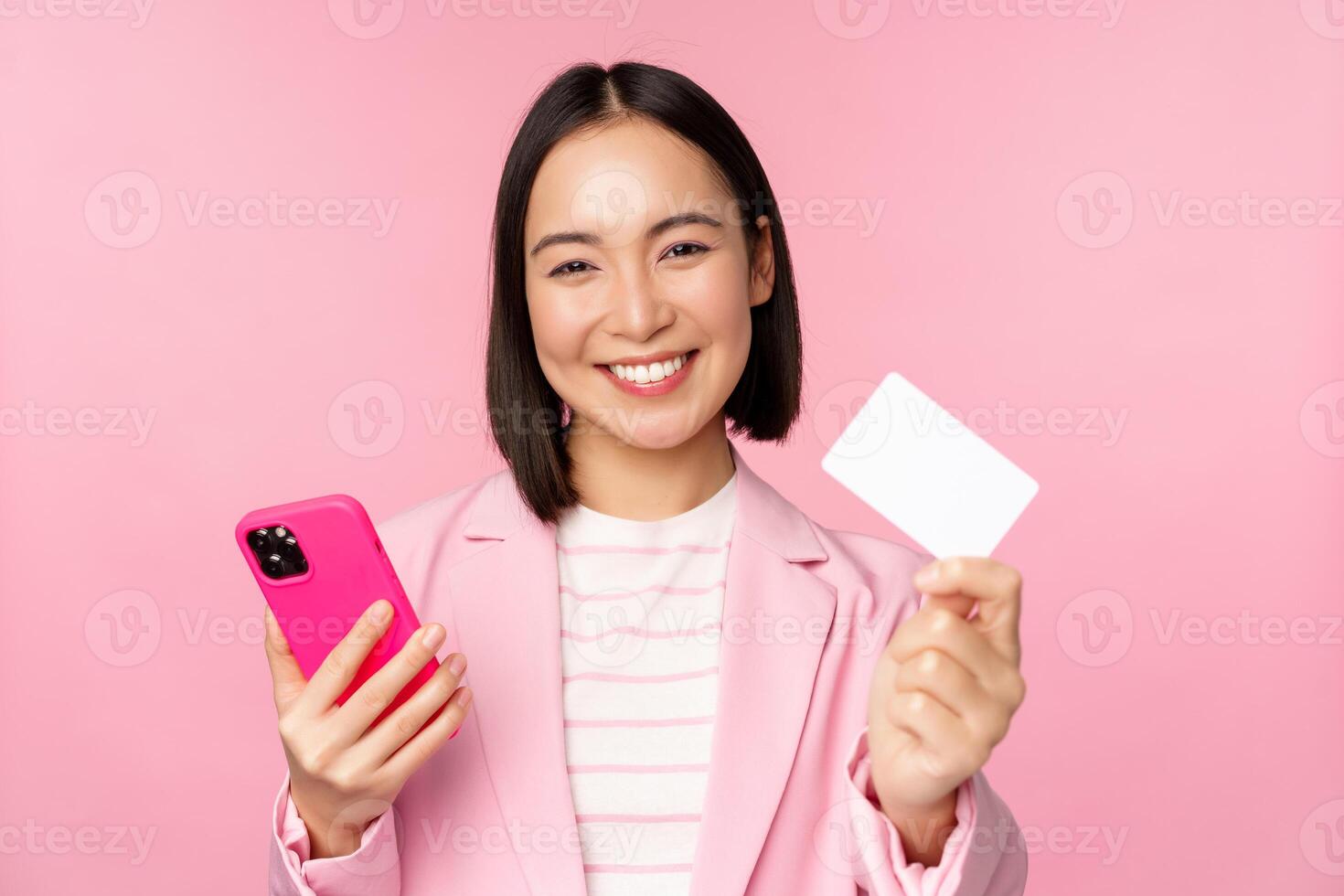 Image of smiling happy asian businesswoman showing credit card, paying online on smartphone application, order with mobile phon, standing against pink background photo