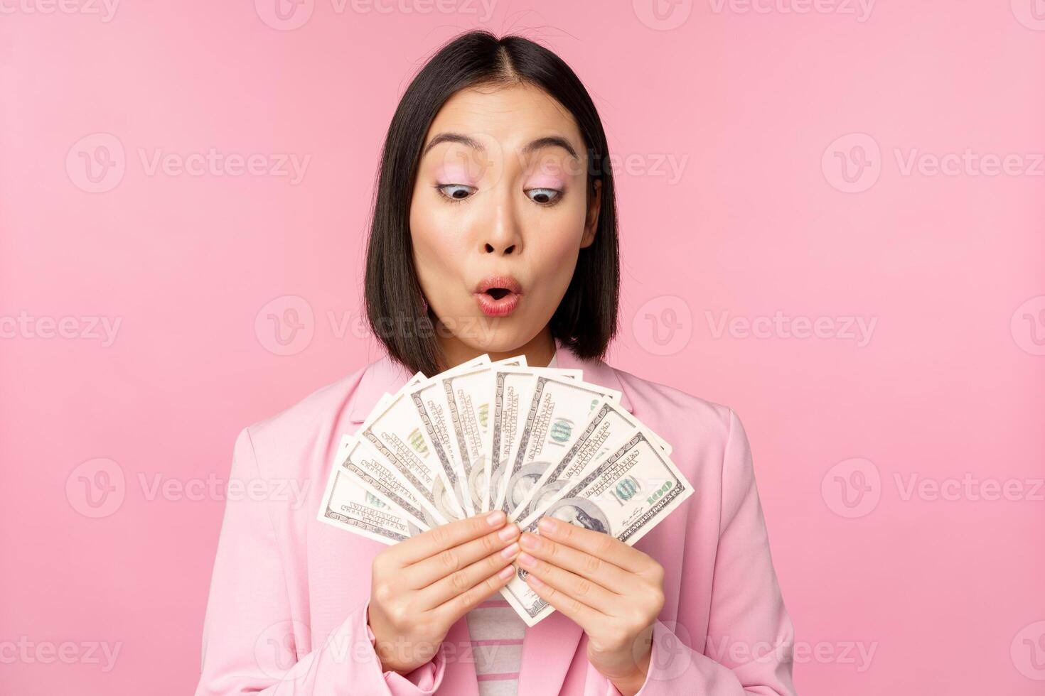 Finance, microcredit and people concept. Happy smiling asian businesswoman showing dollars money, standing in suit against pink background photo