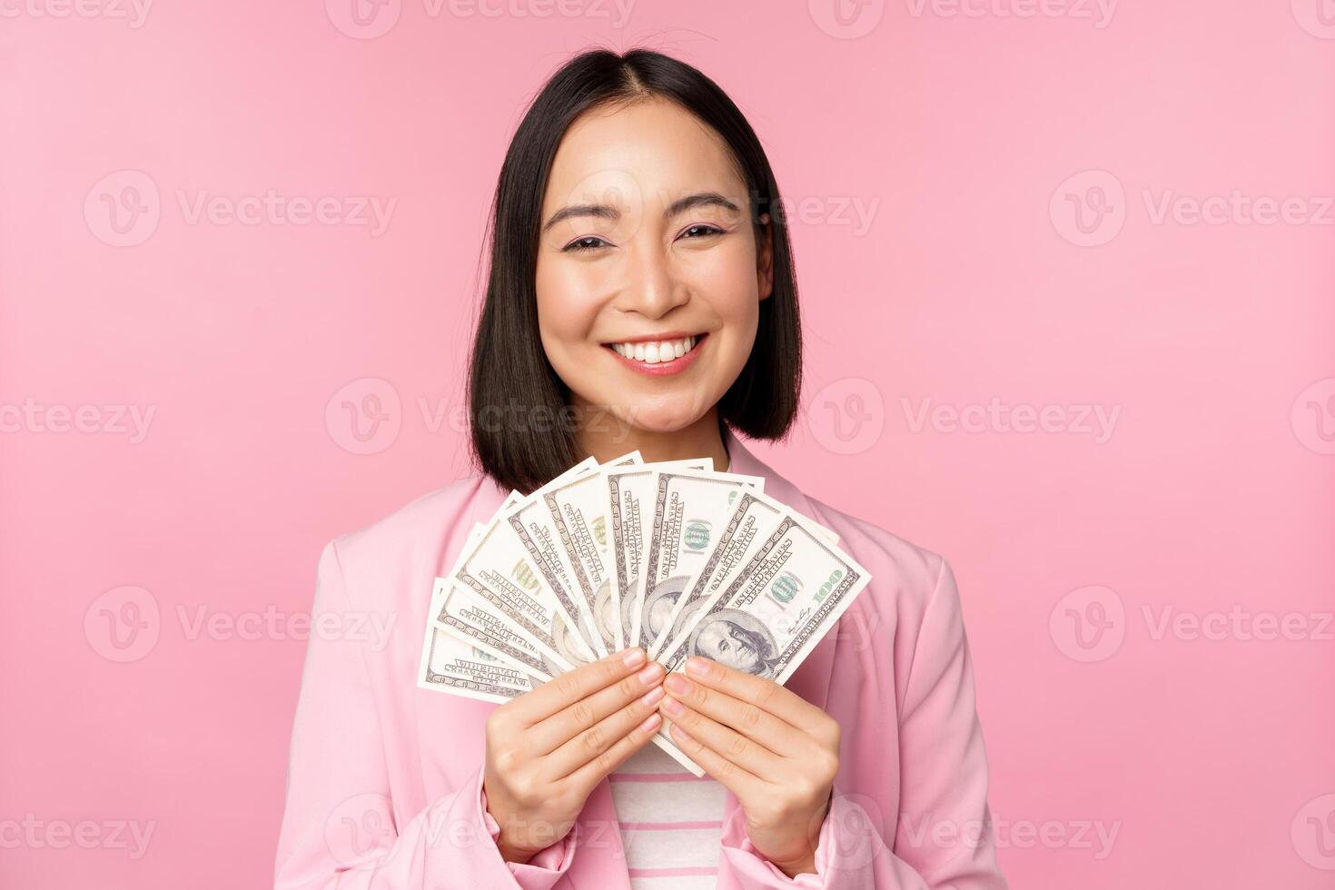 Finance, microcredit and people concept. Happy smiling asian businesswoman showing dollars money, standing in suit against pink background photo