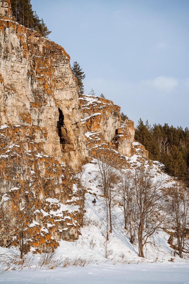 Winter landscape of a rock on the bank of the river. The rocky mountains of orange color are strewn with snow. Cold Russian winter. Ural Mountains photo