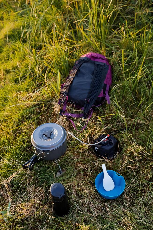 Hiking, outdoor activities. Camping kitchen utensils lie in the grass a thermos, a mug, a heater, a bowl and a backpack. Tourist breakfast. Cooking in nature. photo